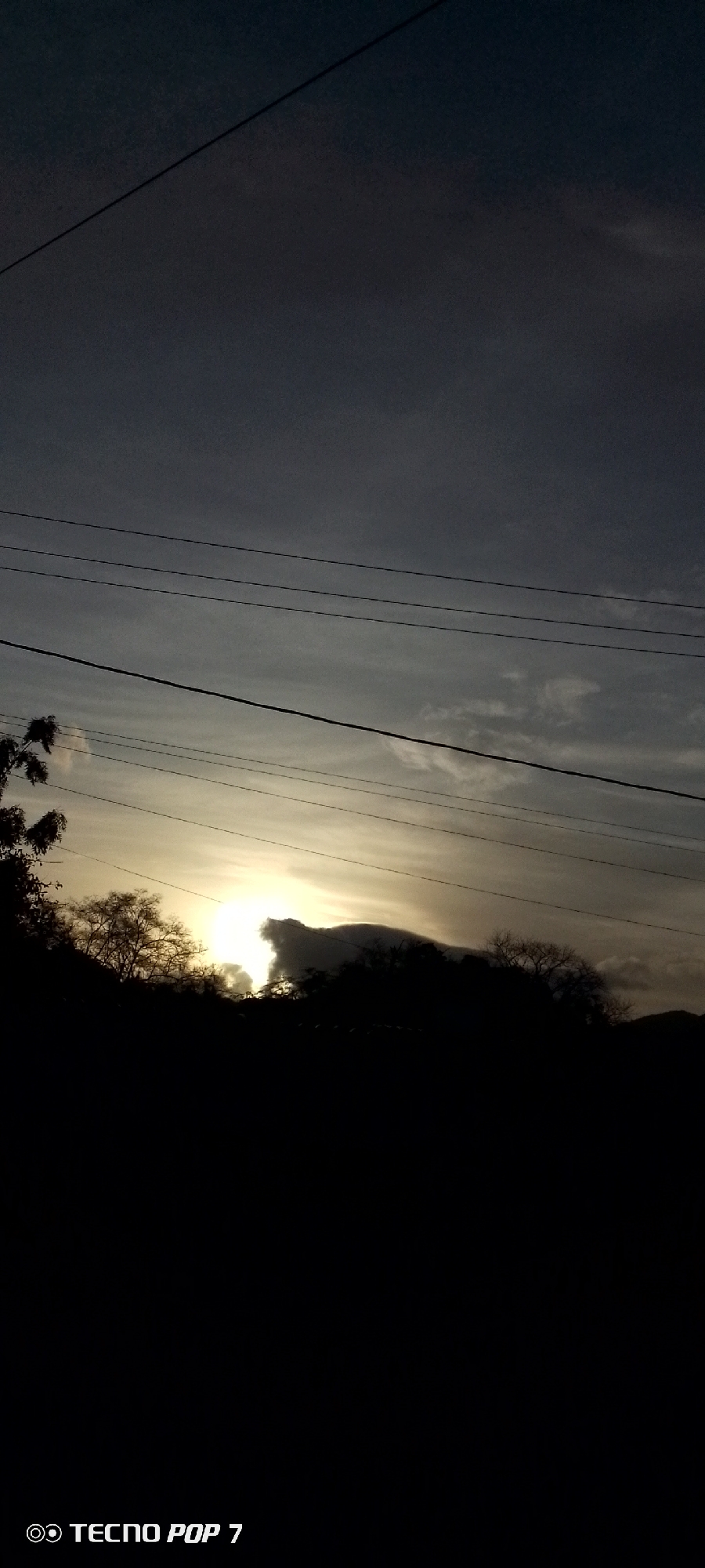 The image depicts a sunset scene in Altagracia, Venezuela. Silhouetted trees and foliage are present in the foreground against a sky transitioning from dark blue at the top to a brighter, sunlit portion near the horizon. The sun is partially obscured by clouds, creating a radiant glow. Electrical wires are strung across the upper portion of the frame. The bottom of the image is almost entirely black. The "TECNO POP 7" watermark is visible at the very bottom.