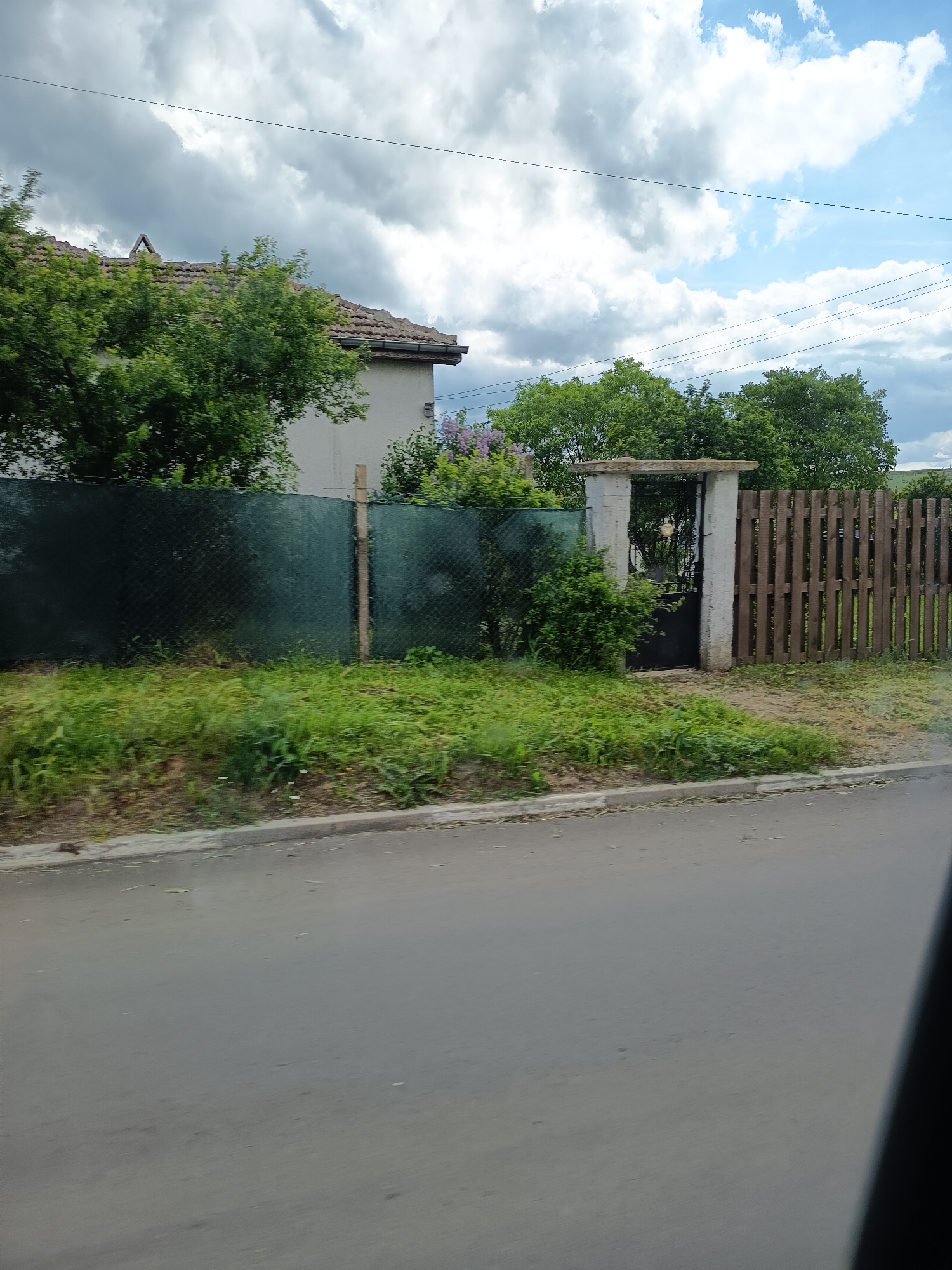 The image presents a suburban scene, likely in Unknown, Unknown. A paved road occupies the foreground, with a curb separating it from a grassy area. Behind the grass, a fence extends across the midground. The fence is composed of two distinct sections: one covered with dark green netting and the other made of vertical wooden slats. A concrete archway with a metal gate provides a passageway through the fence. Vegetation, including bushes and trees, obscures parts of the fence and the building in the background. The building has a tiled roof and light-colored walls, partly hidden by foliage. The sky above is a mix of dark and light clouds, suggesting variable weather conditions. Overhead power lines are visible against the sky. The image appears to be taken from a moving vehicle.
