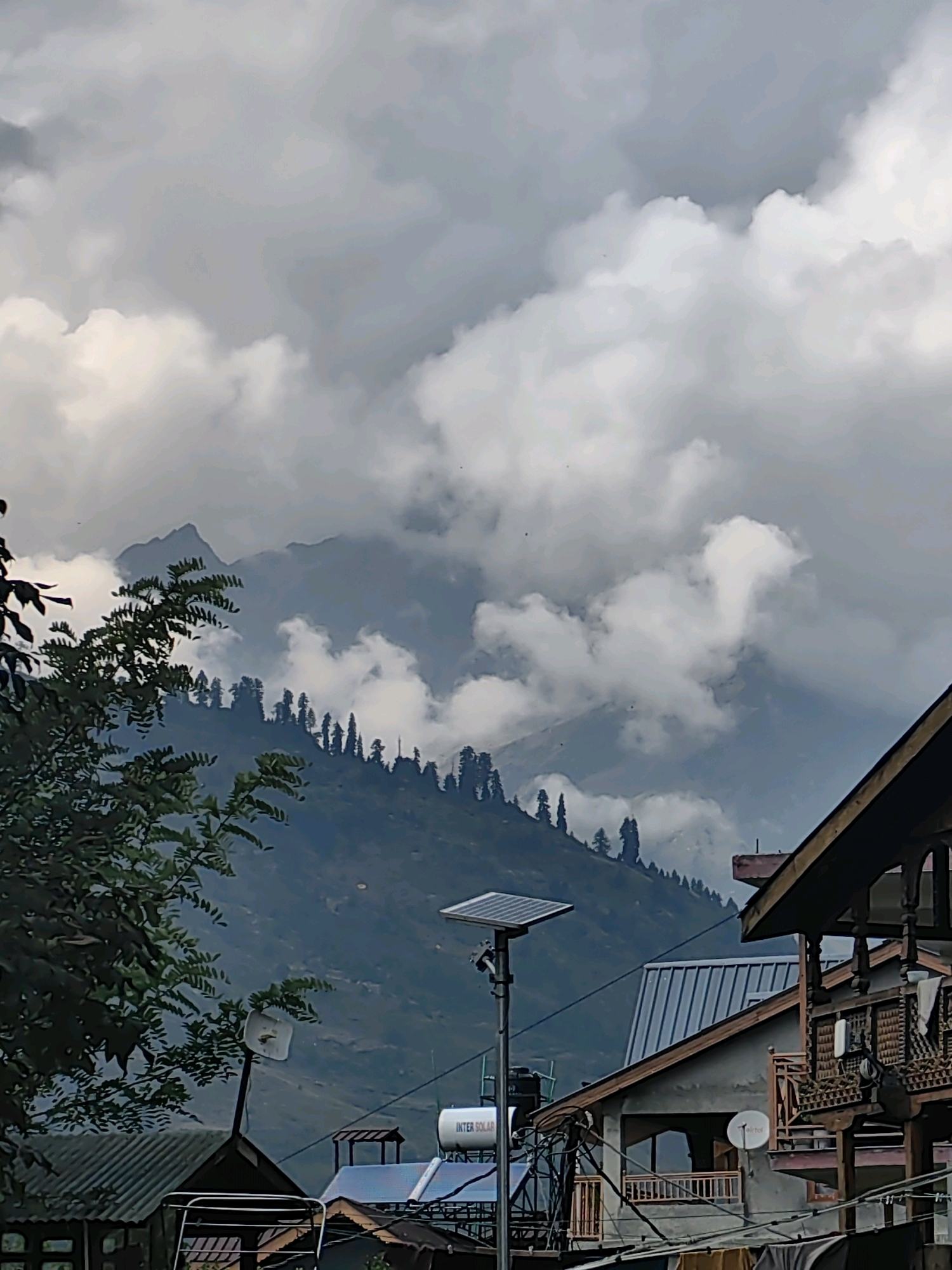 The image depicts a mountainous landscape with a village in the foreground, set in Vashisht, India. The sky is overcast with dramatic, billowy clouds that obscure parts of the mountain peaks. The mountains themselves are covered in lush green trees, primarily pine trees, that form a dense forest line along the slopes.
In the foreground, several buildings are visible. On the right, a traditional wooden house with intricate carvings on its balconies and railings stands out. To its left, a more modern structure with a corrugated metal roof is situated. Further to the left, a house with a sloped roof and a satellite dish on its side is present, along with a solar panel on a tall pole and a water tank labeled "INTER SOLAR." A large tree with green leaves partially obstructs the view on the far left of the image. The overall scene suggests a cool, possibly damp, day in a mountain village. There are no people visible in the image.