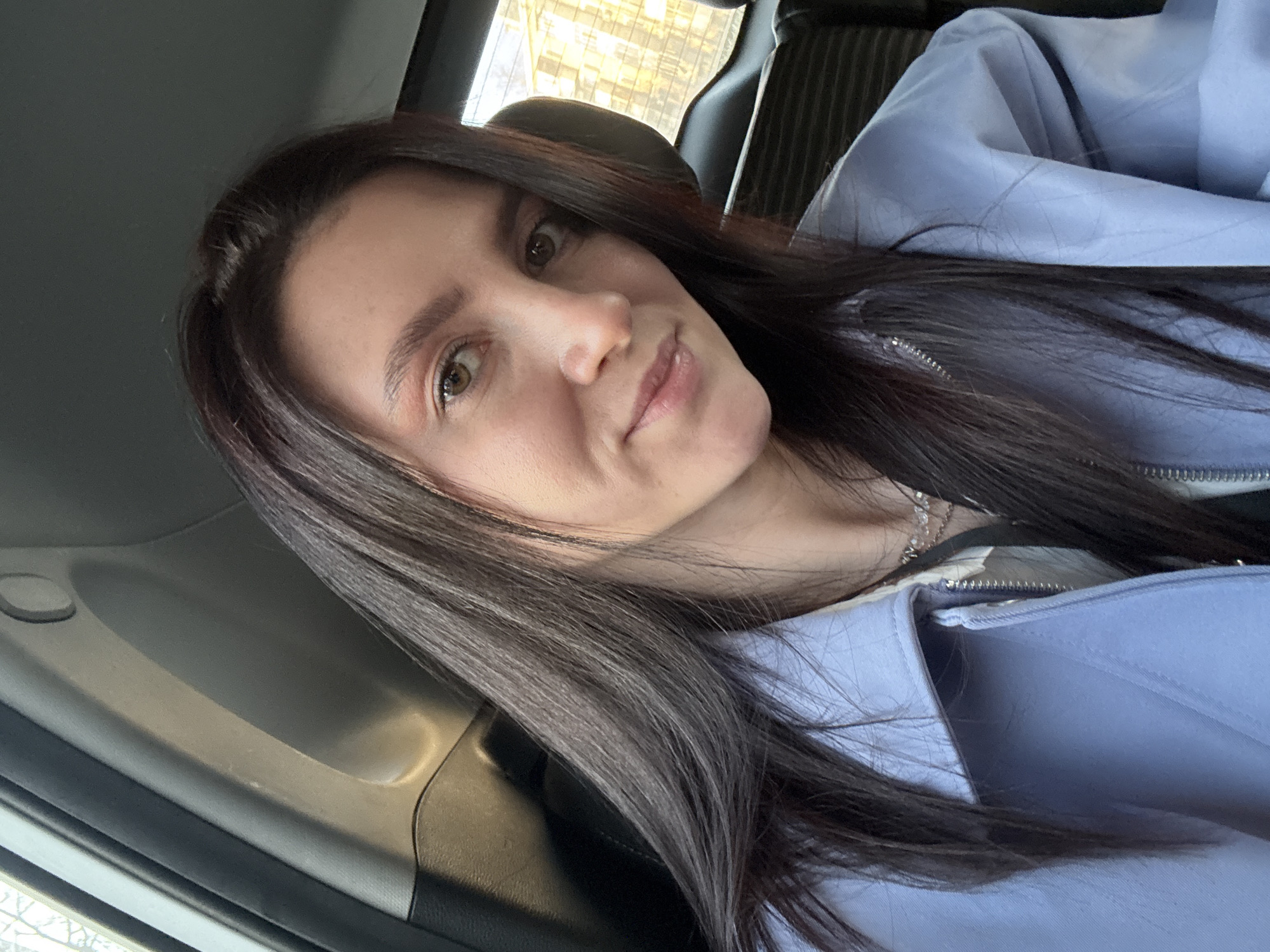 A woman is captured in a selfie taken inside a car in Kazan, Russia. She is positioned in the foreground, looking directly at the camera with a calm and subtly smiling expression. She has long, dark brown hair, olive skin, and light brown or hazel eyes. She is wearing a light blue or lavender jacket, partially unzipped to reveal a lighter-colored garment and a silver chain necklace underneath.
The setting is a car interior, with a dark car seat headrest visible behind her and a grey door panel to the left. Through the car window in the background, a modern building with numerous reflective windows is visible, indicating an urban environment. The lighting suggests it is daytime, with bright natural light entering the vehicle. No visible text is present in the image.