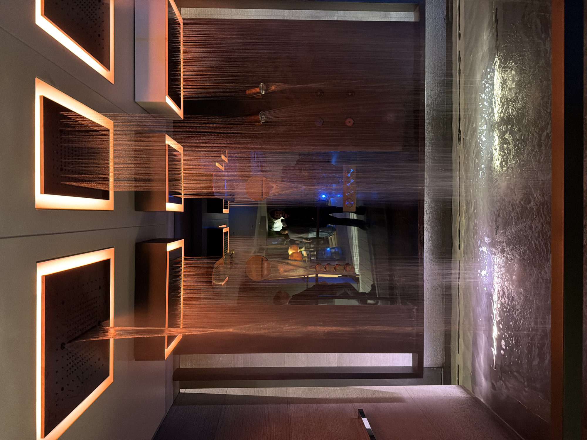 This image depicts an artistic display of water features. On the left, square structures with illuminated borders emit streams of water. In the center, a horizontal plane of water is interspersed with circular elements. On the right, a wall of water cascades downwards. The reflection of individuals and objects is visible in the central water plane. The flooring is made of flat light-colored slabs. The location appears to be an indoor exhibit.