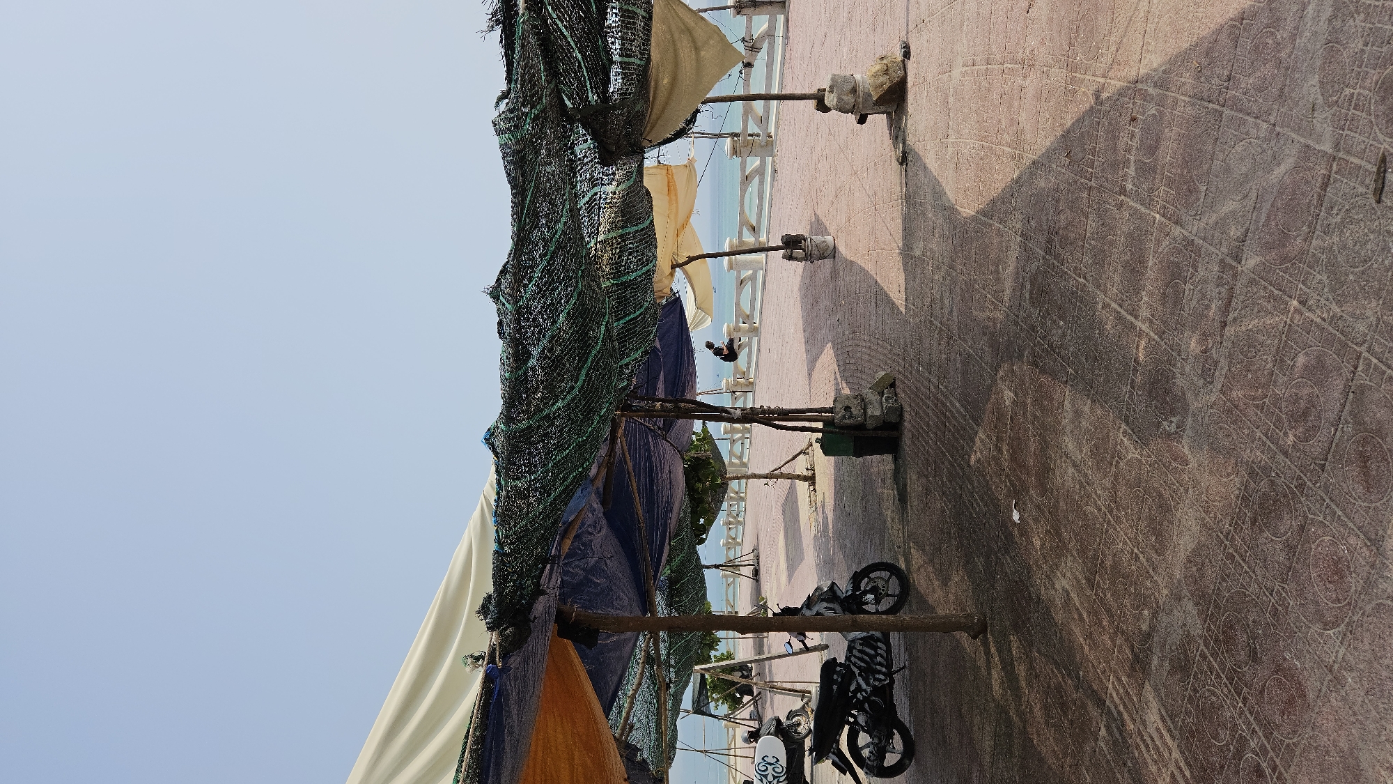 This image depicts a sunny coastal scene in Bac Cam Ranh, Vietnam, along a paved promenade overlooking the sea. The foreground and mid-ground are dominated by several makeshift shelters or stalls constructed from wooden poles, covered with various materials including white, cream, blue, and black netting or fabric tarpaulins.
The ground is a brick-red paved surface, intricately patterned with a grid of intersecting lines and circles. Strong, defined shadows from the overhead structures and poles stretch across this patterned pavement, indicating bright daylight. Two motorcycles are parked on this surface; one is predominantly white, and the other is black.
Beyond the temporary structures, a white concrete balustrade or railing separates the walkway from the calm, blue body of water, which appears to be the ocean or sea. In the distance, between the structures and above the railing, a lone individual can be seen, possibly sitting on or near the balustrade. The sky above is clear and light blue, suggesting good weather.
The scene conveys a tranquil, outdoor setting, possibly a local market, rest area, or beachside vendor stalls. The structures suggest protection from the sun, and the parked motorcycles imply visitors or local activity. No explicit text is visible in the image.
