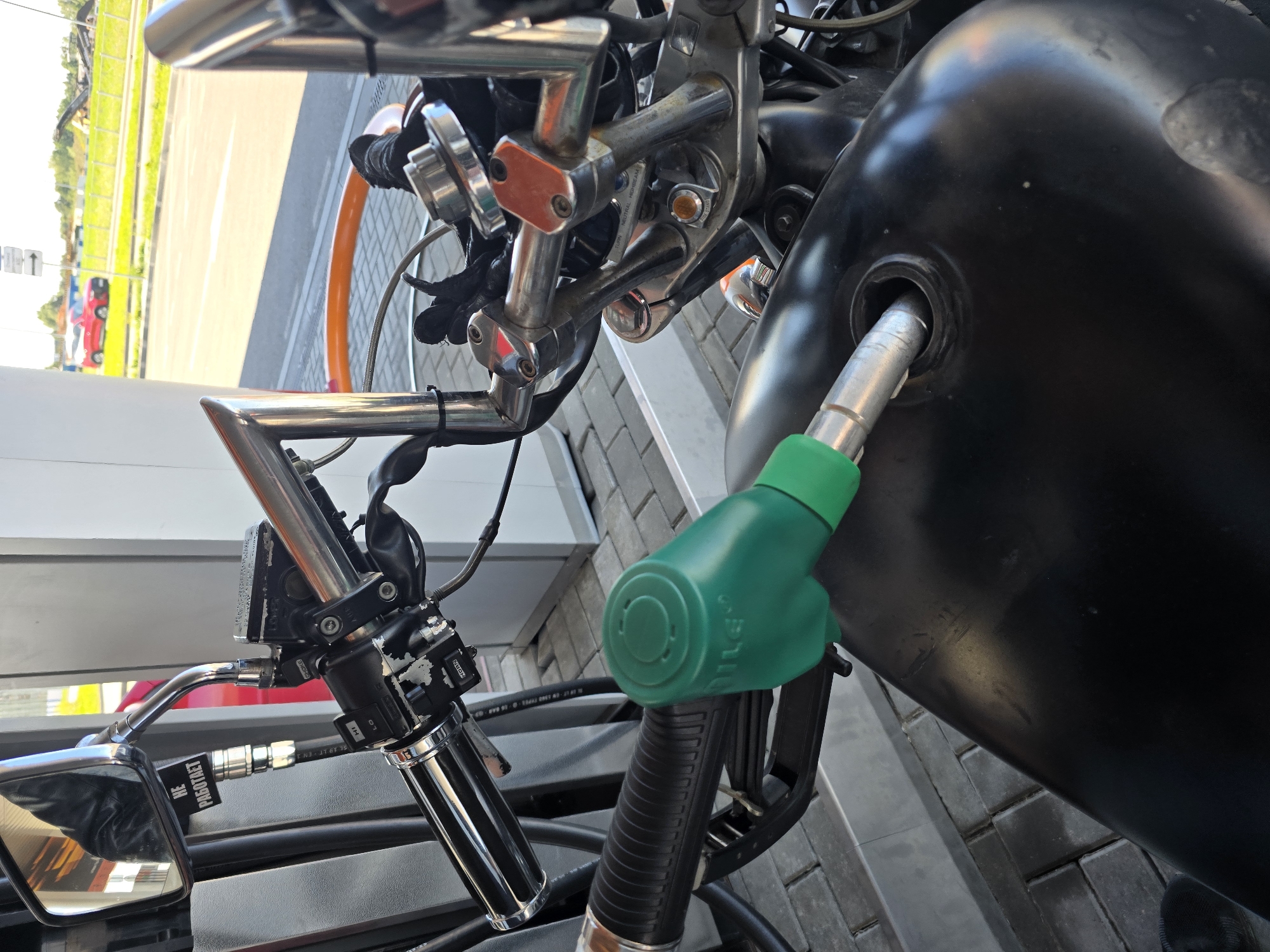 In Холмогоровка, Russia, a close-up, eye-level shot captures a motorcycle being refueled at a gas station. A green fuel nozzle is inserted into the motorcycle's black fuel tank. The handlebars, chrome accents, and part of the motorcycle's front are visible, along with a sticker on the mirror that reads "НЕ РАБОТАЕТ" (does not work). The background shows a paved area and a street with a red car driving by, indicating an outdoor urban setting on a sunny day.
