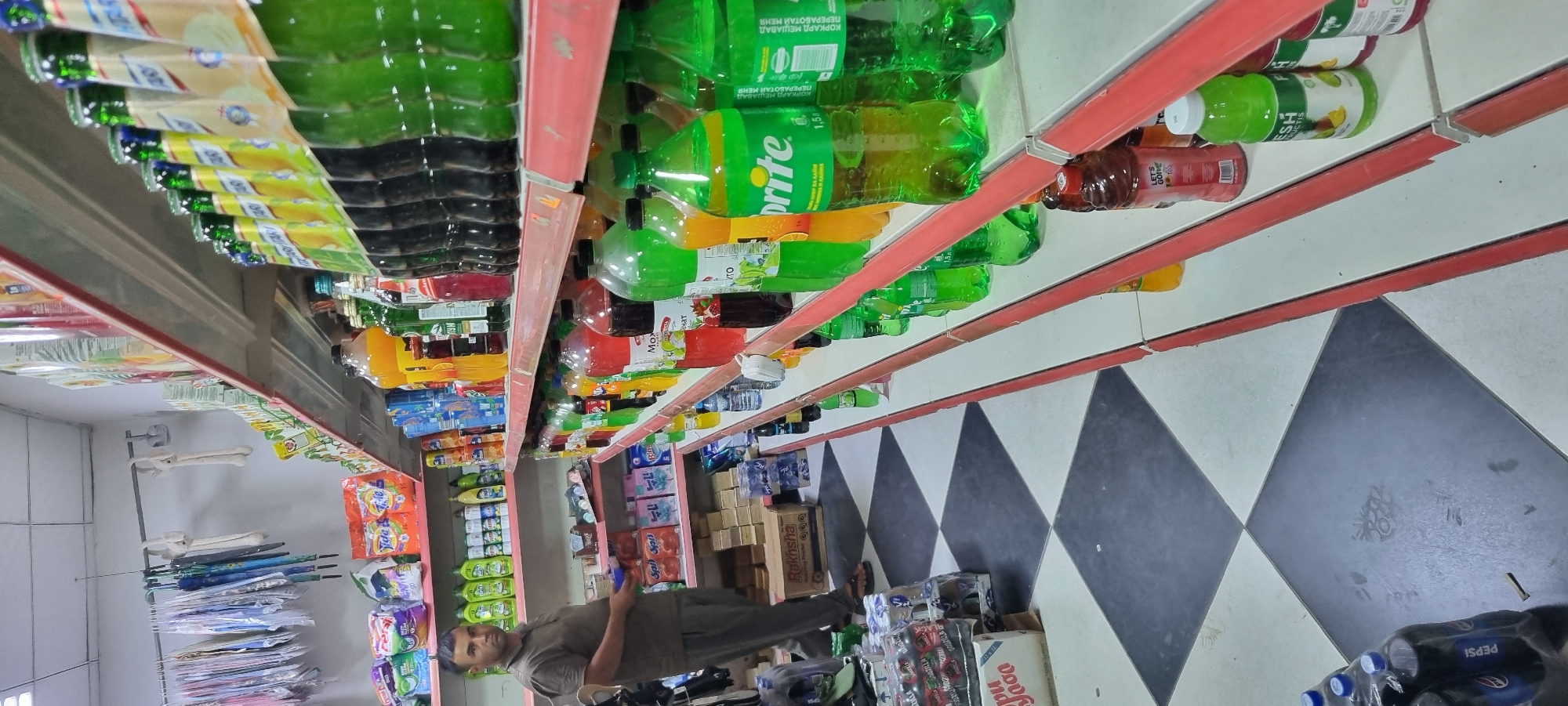 The image depicts the interior of a convenience store, likely in Ismoili Somoni, Tajikistan. A man in a brown t-shirt and grey pants is standing on the left side of the frame, holding a blue tray. He appears to be a store employee or customer. The store is stocked with various items, including cleaning supplies, packaged foods, and a wide assortment of bottled beverages displayed on tiered shelving. The floor has a black and white checkered tile pattern. The shelves are filled with numerous plastic bottles of soft drinks and juices, many of which are green and yellow. The lighting appears to be artificial, typical of an indoor retail environment.