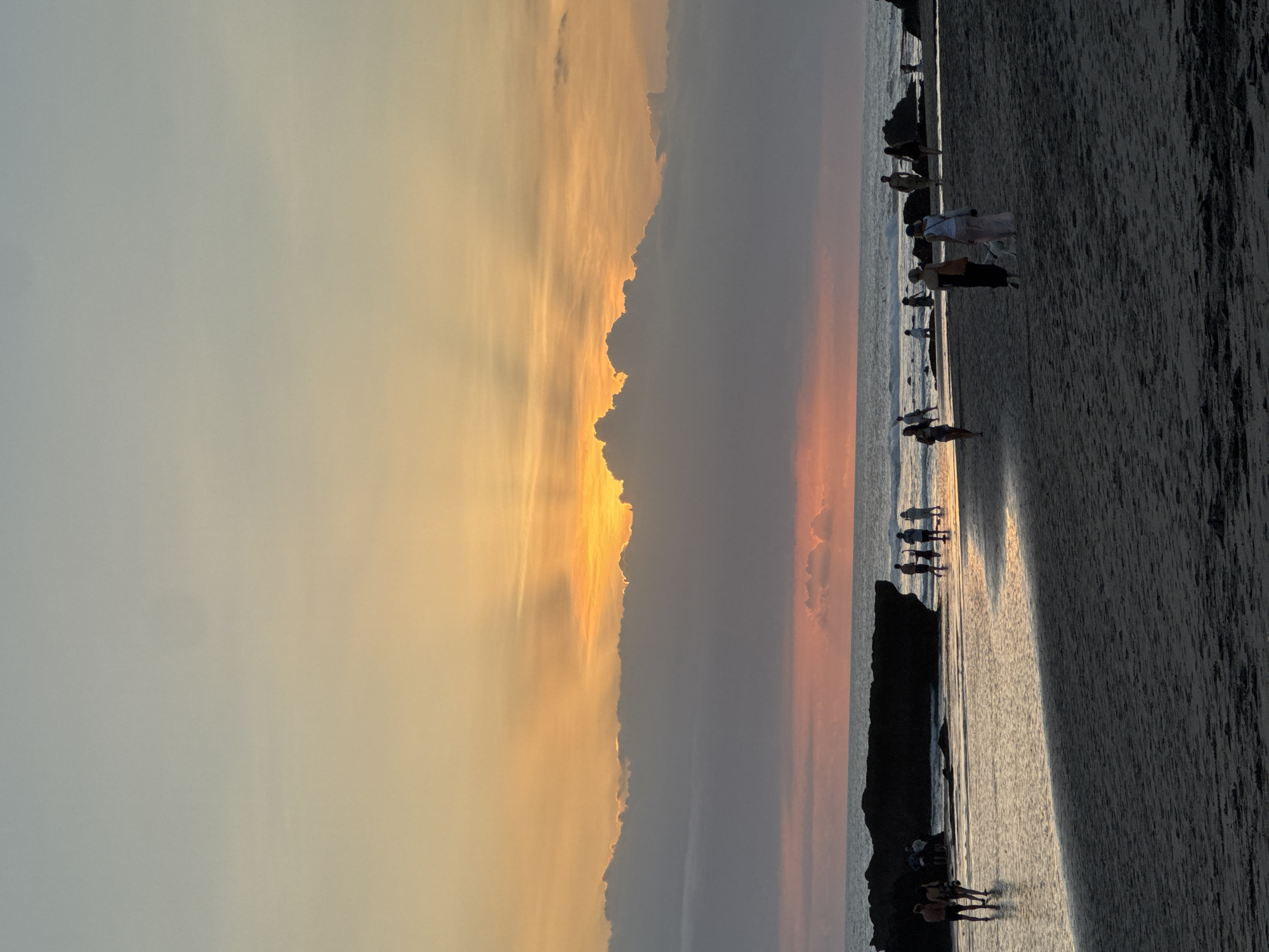 A vertical, medium shot captures a beach scene, likely in North Kuta, Indonesia, during sunset. The sky transitions from light blue to orange hues, with some clouds present. A line of darker, ominous clouds sits between the sky and the ocean. On the beach, there are several small groups of people, mostly standing and appearing to watch the sunset. The ocean stretches from the shore towards the right side of the frame.