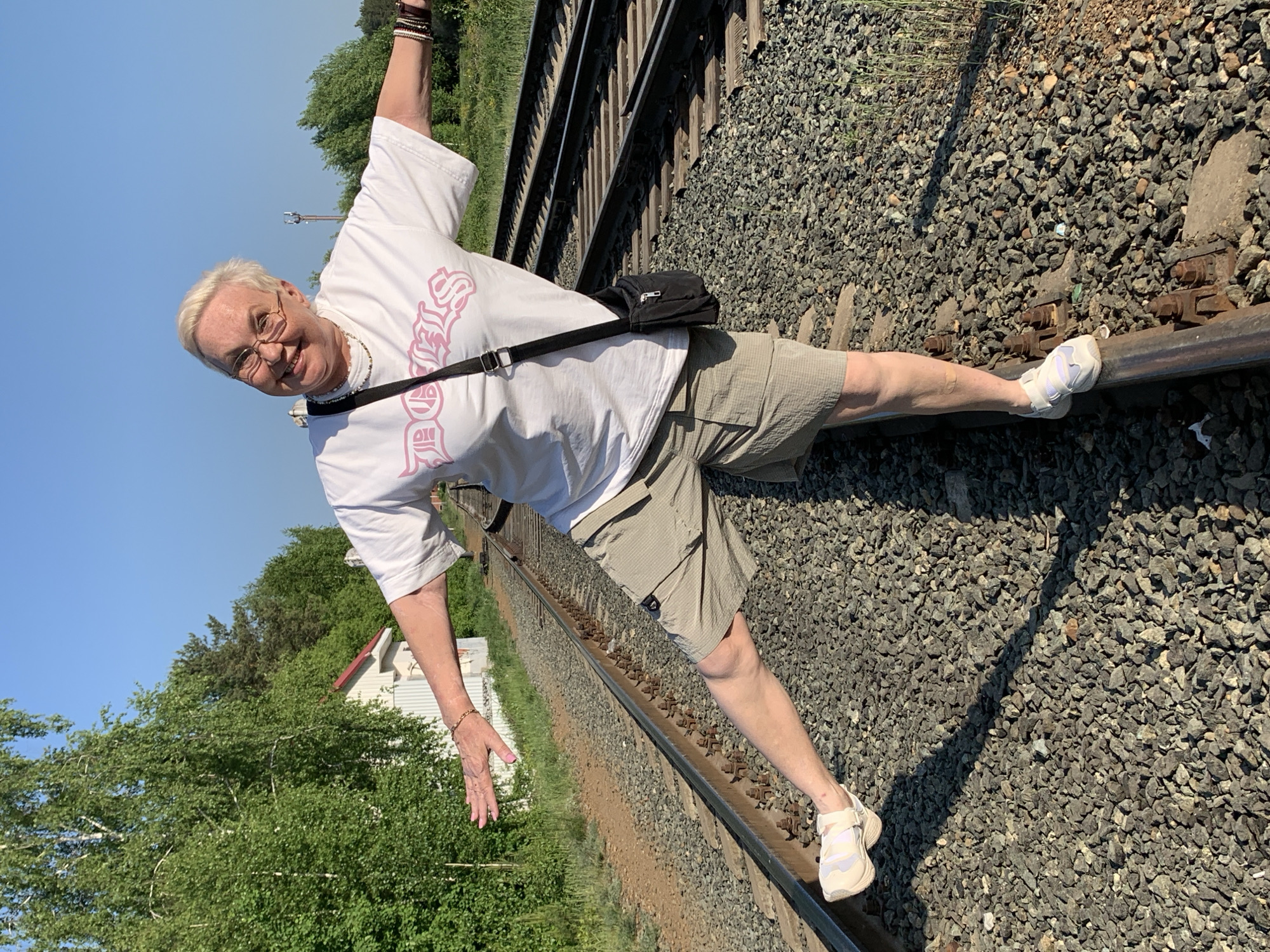 A mature woman stands on a railroad track in Chaikovsky, Russia. The railroad track is elevated and covered in gravel. She balances with one foot on the rail, while the other leg is extended straight, resting on the opposite rail. She wears a white T-shirt with pink text, khaki shorts, and white sneakers. A black shoulder bag hangs across her body. She has short blonde hair, glasses, and is smiling. One arm is raised overhead, the other extended to the side for balance. Trees and a white building with a red roof are visible in the background. The sky is clear and blue.