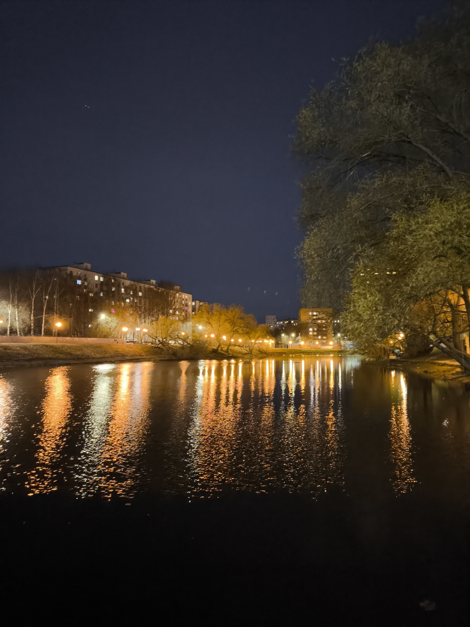This nighttime photograph captures an urban landscape, likely in Khimki, Russia, centered around a tranquil body of water. The upper portion of the image features a dark, deep blue sky, with a few faint specks of light possibly indicating distant stars or artificial illumination.
Along the far bank, a series of multi-story buildings, characteristic of residential architecture, are visible, with numerous windows emitting light. The banks of the water body are lined with trees; those on the left appear to be bare or in early foliage, while a large tree on the right, adorned with green leaves, partially frames the scene.
The most striking feature is the reflection of artificial lights on the water's surface. Numerous streetlights and building lights cast a warm, golden-orange and white glow, which is spectacularly mirrored. These reflections are elongated and fragmented by the gentle ripples in the water, creating vibrant, shimmering streaks of color that extend across the lower half of the frame. The immediate foreground of the water remains dark, transitioning into these bright patterns.
The image portrays a peaceful and quiet urban evening. There is no discernible human activity taking place, nor any visible text within the frame.