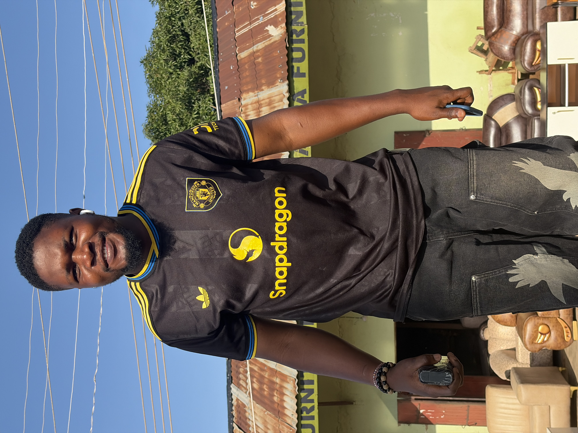 A Black man is standing outside a building, with a clear sky in the background. He is wearing a black Manchester United football jersey with yellow and blue trim, and dark washed denim jeans with a bird graphic on the right leg. He has a beard and is smiling at the camera. He is wearing white wireless earbuds. In his left hand, he is holding a dark colored object, possibly a phone or a power bank, and in his right hand, he is holding a blue phone. Behind him, the building has a corrugated metal roof and appears to be a furniture shop, with various sofas and chairs visible both inside and outside. The text "FURNI" is visible on the building, suggesting it is a furniture store. The location is identified as Gura Top, Nigeria.