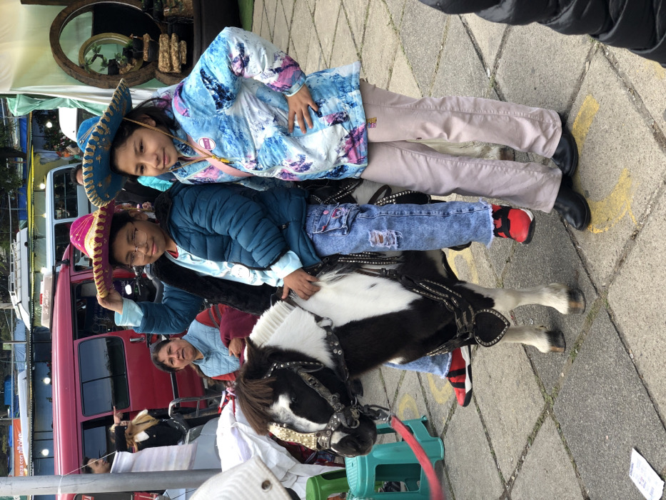 Two young girls in sombreros are on a black-and-white pony. One wears a blue jacket and ripped jeans with red-and-black sneakers, while the other sports a blue, tie-dye style coat and tan pants with black shoes. They pose atop the pony. An adult woman stands nearby. A pink bus is visible behind them.
