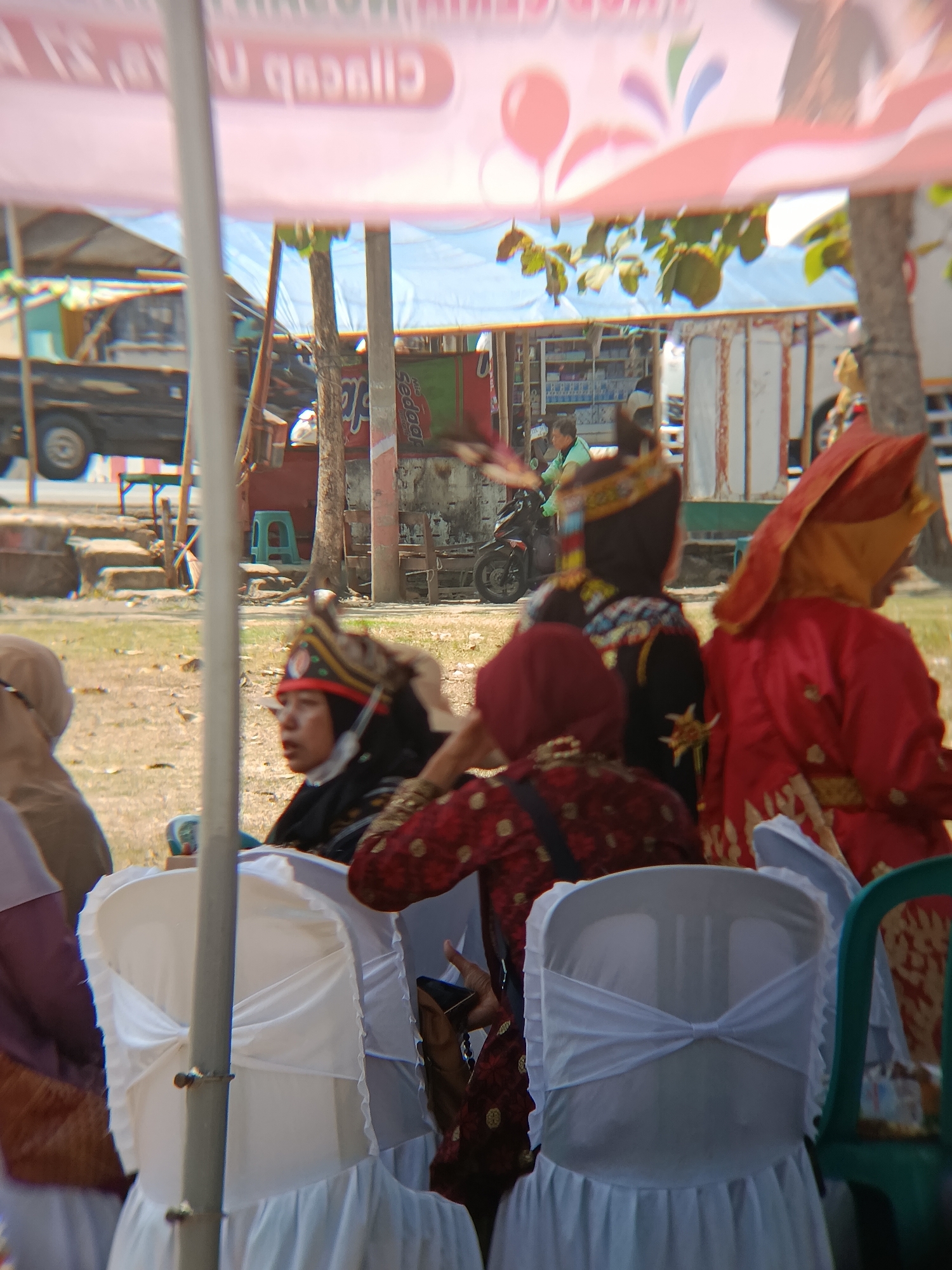 The image depicts an outdoor scene in Desa Kebon Manis, Kecamatan Cilacap Utara, Indonesia, likely a cultural event or gathering. In the foreground, several chairs are draped with white fabric, creating a formal seating arrangement. Some individuals are seated, observing.
Several people are visible, dressed in traditional attire. One woman in the center foreground wears a red and black patterned garment and a distinctive headdress with red trim and gold accents. To her right, another person is seen from behind, wearing a vibrant red traditional outfit with yellow accents and a matching headscarf. Further to the left, a person in a light brown hijab is partially visible.
In the background, the scene transitions to a street or open area. A dark-colored pickup truck is parked. Beyond that, there appears to be a market stall or shop with shelves displaying items. A motorcycle is also visible in the mid-ground. The overall environment suggests a sunny day, with a canopy or tent providing shade over the seating area. The blurry nature of the background indicates a shallow depth of field, focusing attention on the foreground subjects. The presence of traditional clothing and the general atmosphere suggest a local celebration or cultural performance.