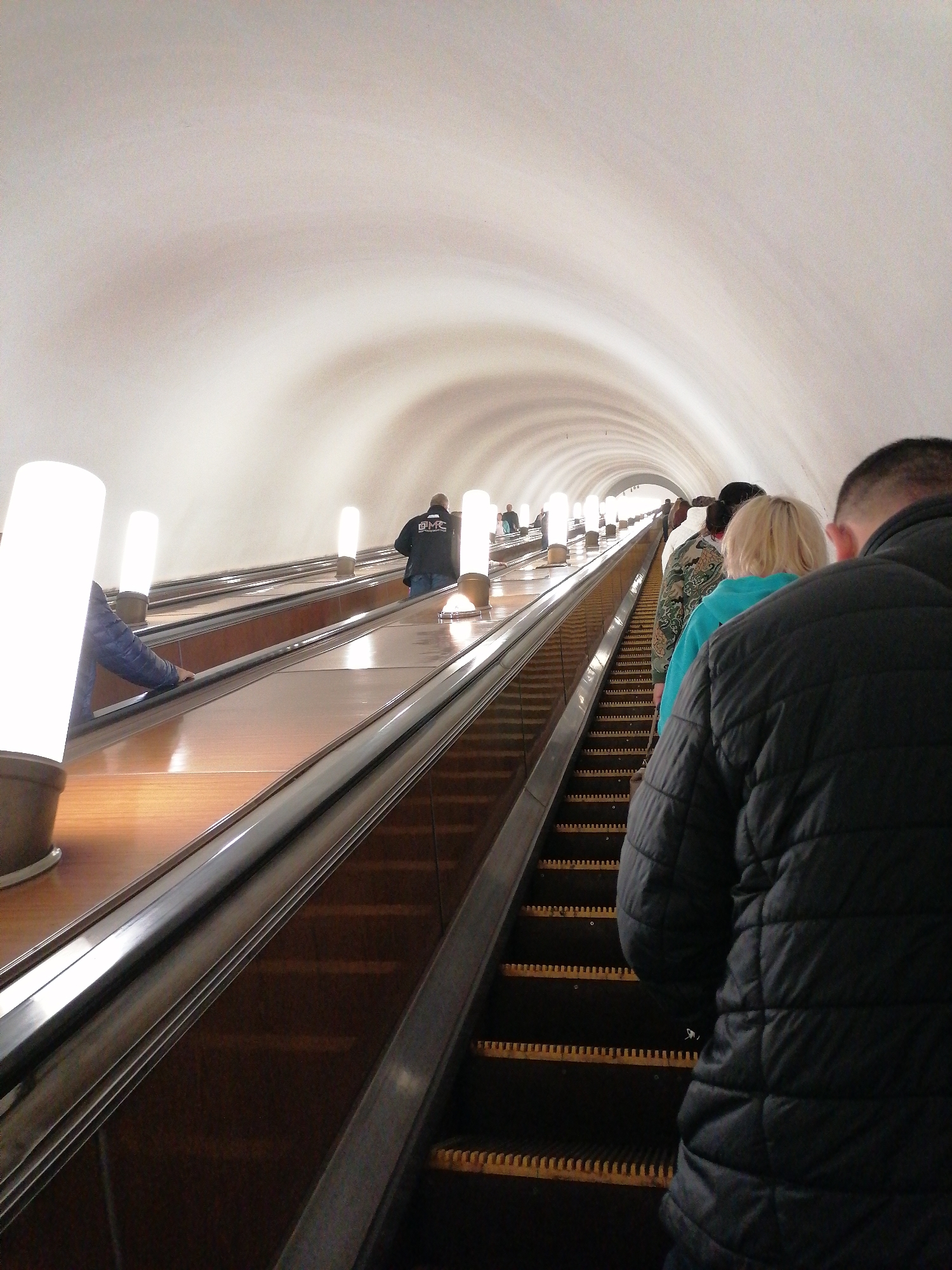 The image depicts a deep underground escalator tunnel, likely in a metro or subway station, filled with people ascending. The perspective is from behind a person in the foreground, looking up the long incline.
Several individuals are visible on the escalators. The closest person is seen from the back, wearing a black quilted puffer jacket. In front of them, a person with blonde hair wears a light blue top, followed by another in a camouflage-patterned jacket. On a parallel escalator to the left, a person in a blue jacket has their hand on the moving handrail. Further up, another person on the left escalator wears a dark jacket with a discernible "DMC" logo on the back.
The escalators feature dark steps with yellow comb plates, dark metallic handrails, and glossy wooden-like side panels. Bright, vertical cylindrical white light fixtures are mounted at regular intervals along the escalator sides, illuminating the path. The tunnel ceiling is a light, possibly white or cream, color with a ribbed or corrugated texture, creating a distinct visual pattern that narrows towards the distant top of the escalator. The environment is entirely artificial, characteristic of a subterranean transport system, with no indications of natural light or external weather. The scene conveys a sense of passive movement during a commute or journey.