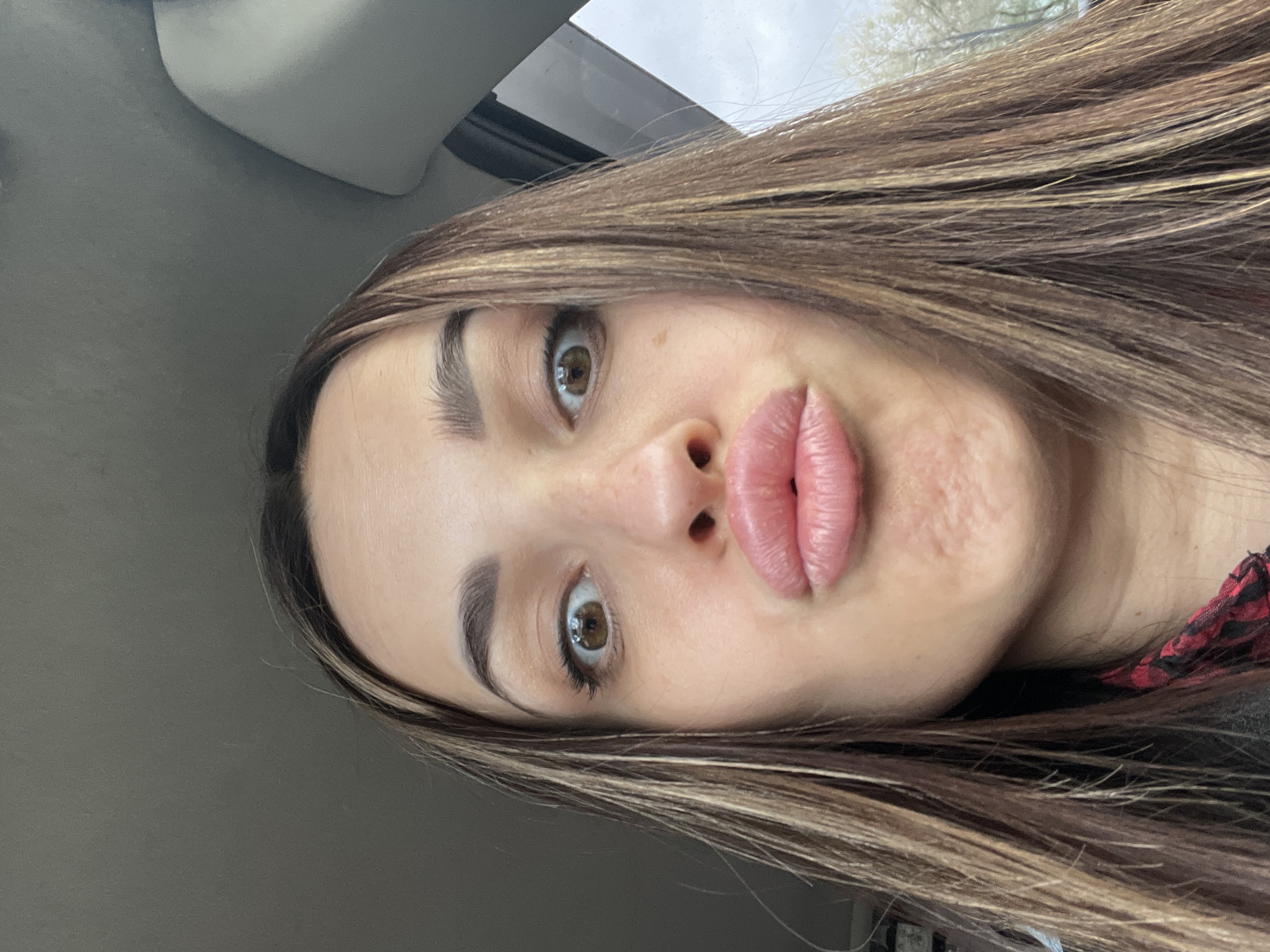 A close-up selfie of a young woman with brown hair and brown eyes. She has fair skin with some freckles and prominent lips. The background appears to be the interior of a car, judging by the gray upholstery and window. The location in the city of Unknown, Unknown is not discernible.