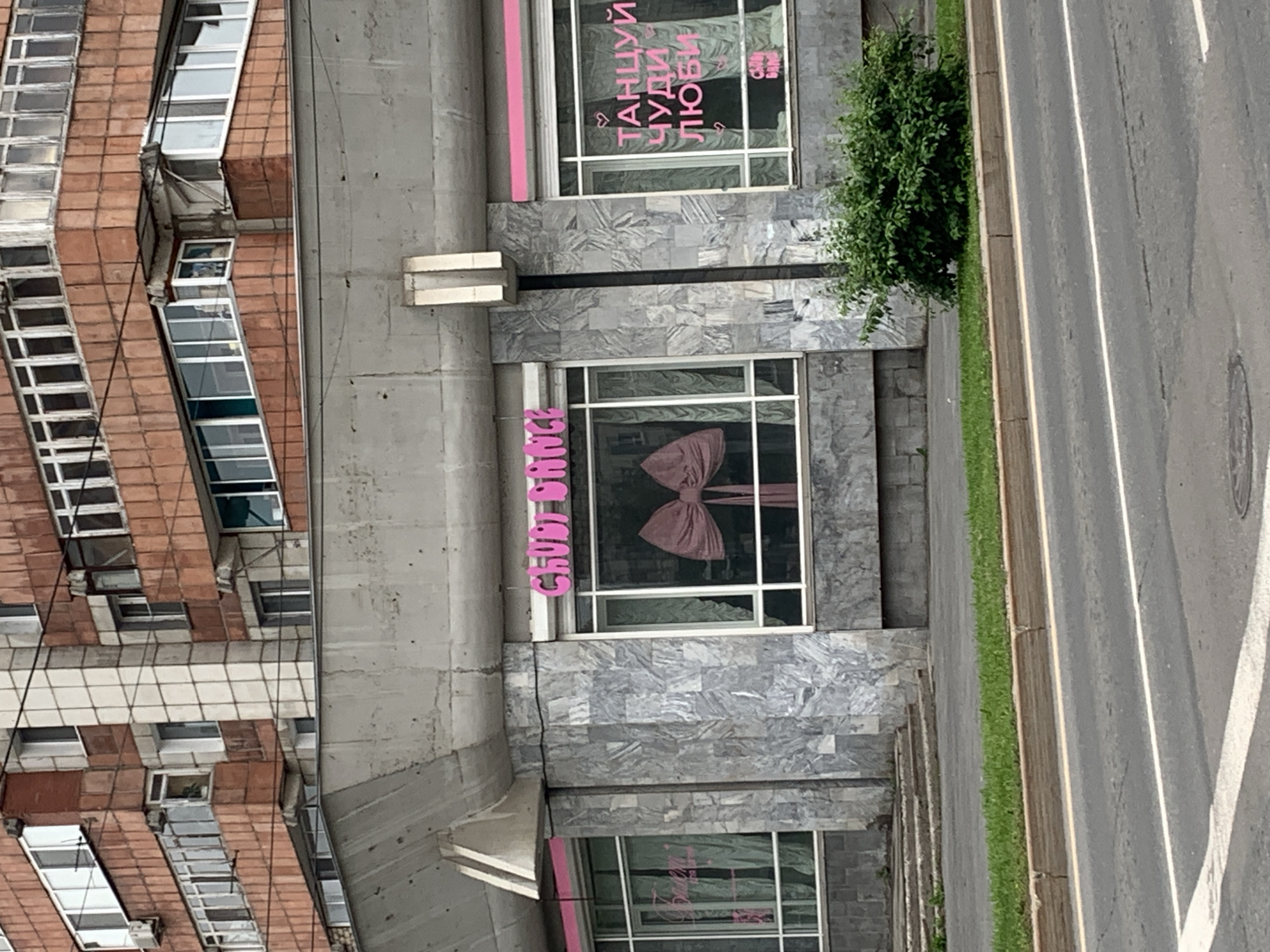 The image captures a storefront on a cloudy day, with a street in the foreground and a multi-story apartment building in the background. The storefront features large windows with pink lettering. On the left window, green curtains and a pink beanbag chair are visible. The central window displays a large, pink bow. The right window has pink lettering and two small heart symbols. The building appears to be made of concrete and marble-like tiles. A small patch of green grass and a shrub are located between the storefront and the street. The apartment building in the background is constructed with red brick and has numerous windows, some of which are enclosed balconies. There are no people visible in the image. Based on the Cyrillic script on the signage, the location is likely Russia, and potentially the city of Perm, though this cannot be definitively confirmed from the image alone.