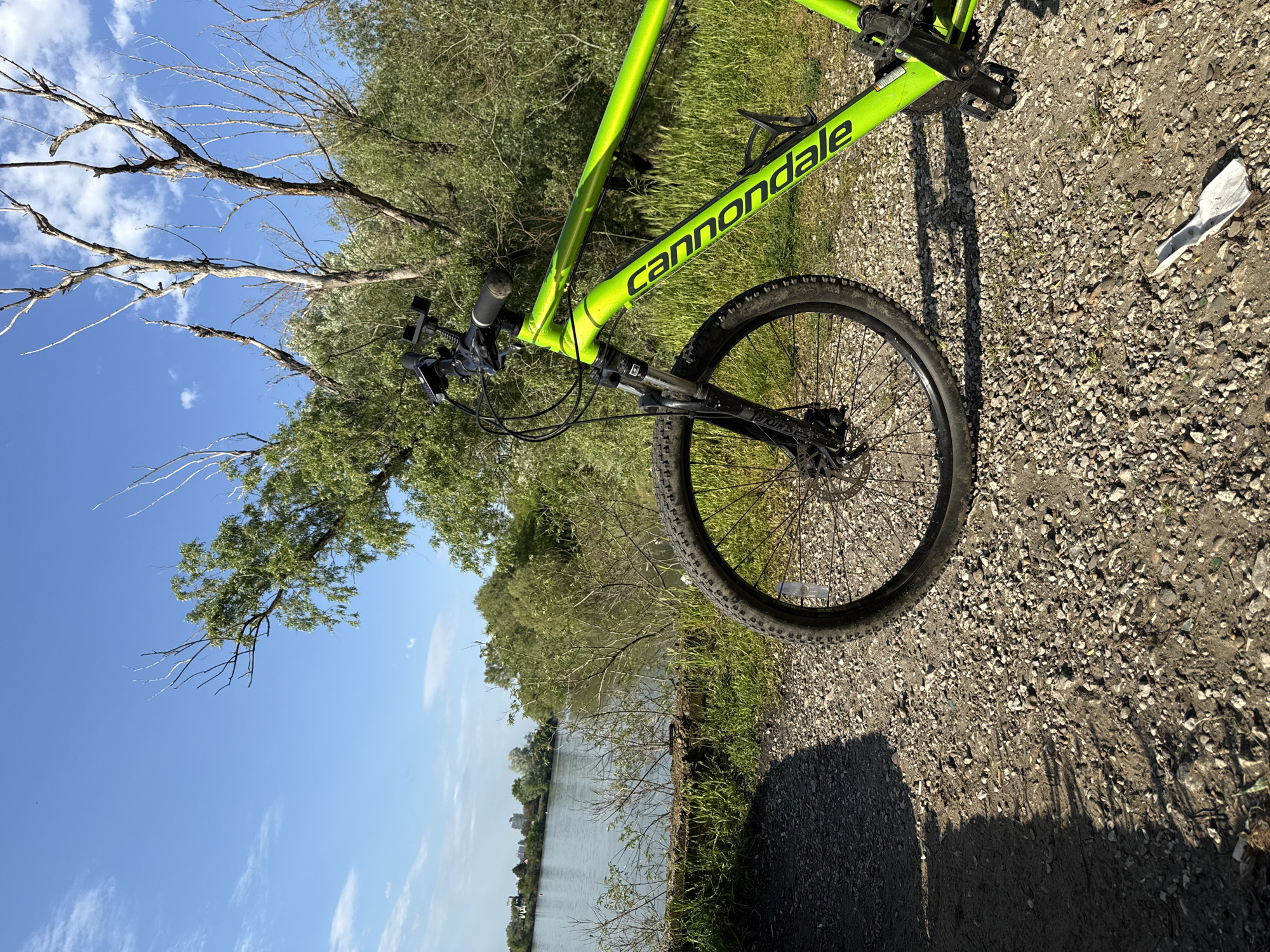 A green Cannondale mountain bike stands on a gravel path alongside a body of water, possibly a lake or river. The background features trees and a blue sky with some clouds. The bike is positioned at an angle in the frame, showing the front wheel, frame, and handlebars. A piece of white paper lies on the gravel path. The city of Unknown, Unknown is not visible in the image.