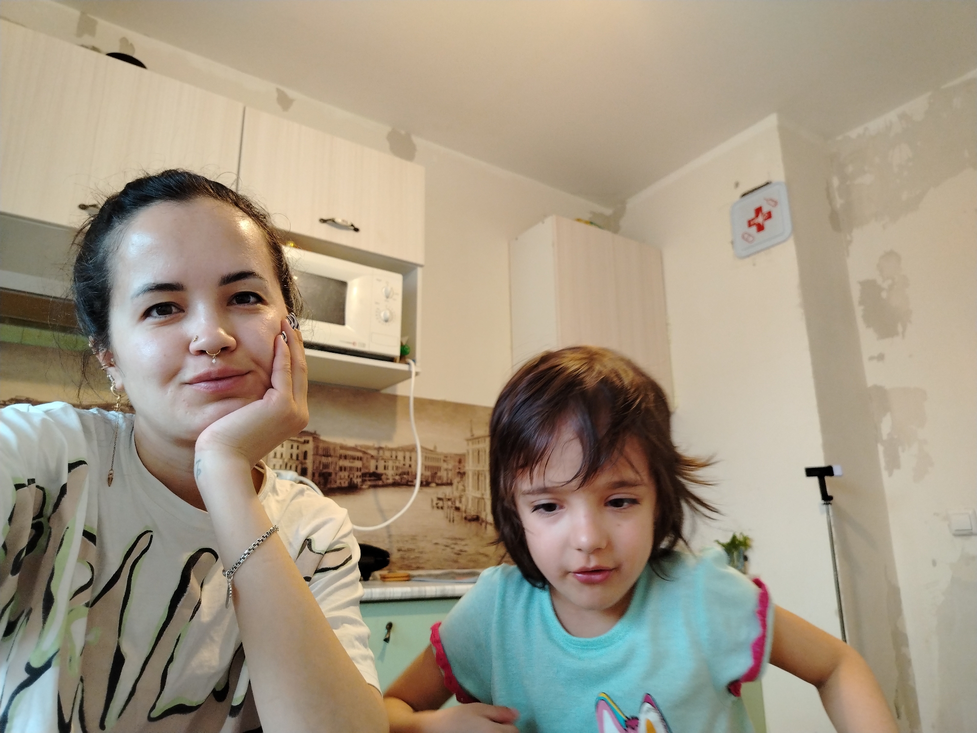 The image shows a woman and a young girl, possibly in a kitchen. The woman, fair-skinned with dark hair pulled back, is wearing a light-colored t-shirt with black and green designs. She has a nose piercing and is resting her chin on her hand. A bracelet is visible on her left wrist.
The girl, fair-skinned with brown hair, is wearing a blue t-shirt with pink ruffled sleeves.
Behind them is kitchen cabinetry, including a microwave. There is also a printed image depicting a city skyline and water, possibly Venice. In the background, a first aid kit can be seen mounted on the wall. The wall has patches of damaged paint.
There is nothing in the image to specifically identify the location as Noviy, Russia.
