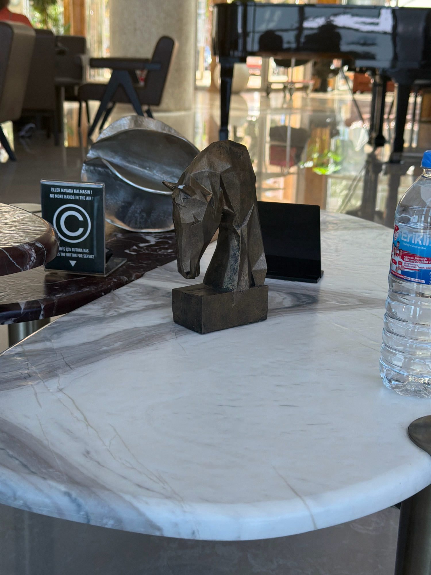 The image depicts a table with a marble top, likely in a hotel lobby or restaurant. On the table, there is a small, stylized metal sculpture of a horse's head, a service call button with the text "ELLER HAVADA KALMASIN! NO MORE HANDS IN THE AIR!" and "RVIS İÇİN BUTONA BAS PRESS THE BUTTON FOR SERVICE", a shiny metal object, and a plastic bottle of water. The background shows seating areas, a grand piano, and large windows reflecting the interior and exterior. The overall setting suggests a comfortable and possibly luxurious establishment. The provided information states the location is in Kuşadası, Turkey.