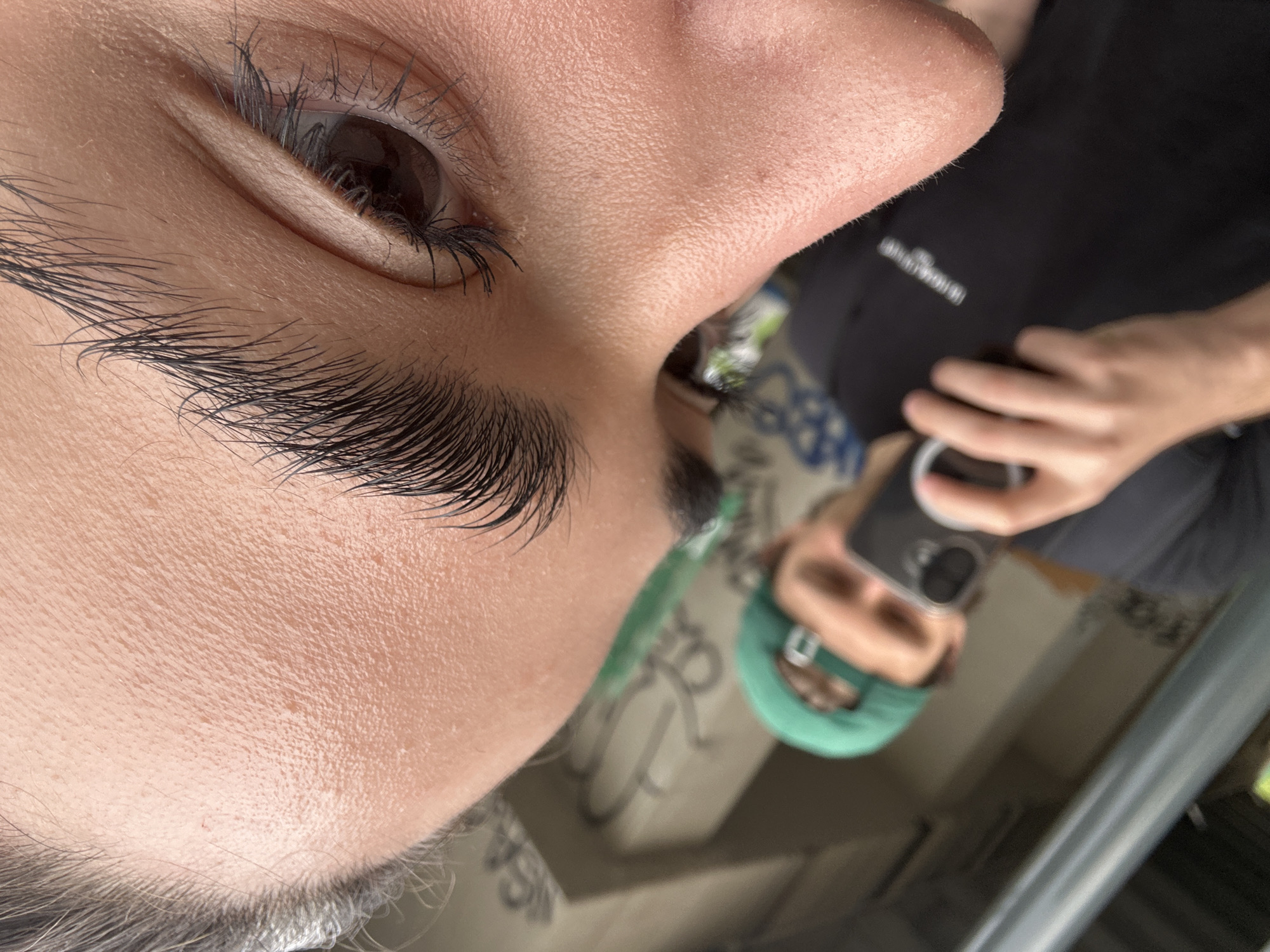 A close-up, upside-down selfie depicts a person with dark eyelashes, a brown eye, and groomed eyebrows. The person is wearing a black top. Reflected in the phone screen is the person's face, a green headband, a beige surface with graffiti-like markings, and a glimpse of an exterior setting with greenery. The specific location within Milan, Italy, cannot be determined from the visual information provided.