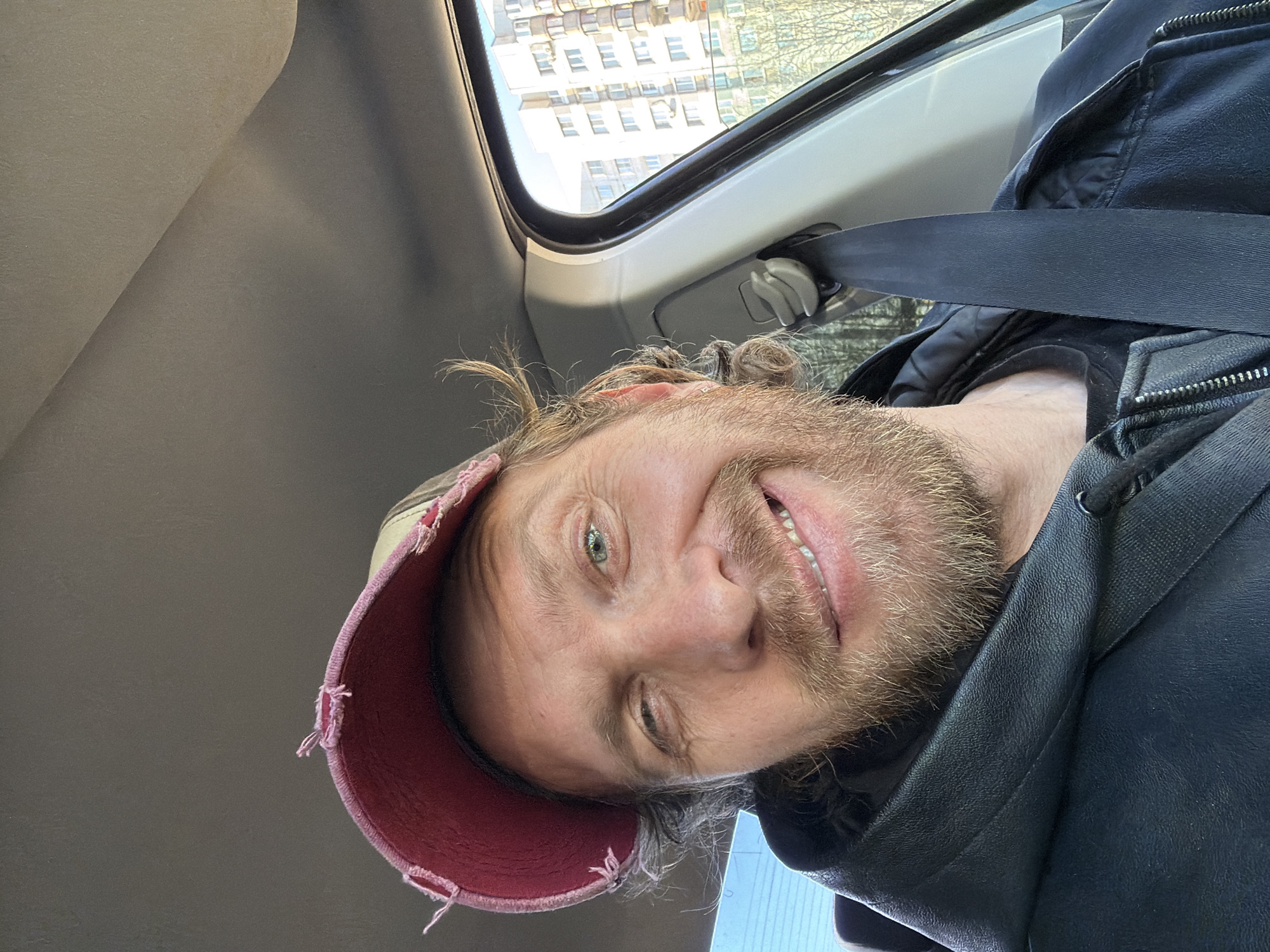 A male individual, likely in his 30s or 40s, is depicted in a close-up selfie taken from inside a vehicle. He has light brown, wavy hair, a full beard and mustache of a similar color, and light-colored eyes. He is smiling broadly, revealing his upper teeth, and winking with his left eye. He wears a distressed baseball cap with a frayed red brim and a lighter-colored crown, along with a dark, possibly black, hooded jacket with a visible zipper. A dark seatbelt is secured across his right shoulder and chest. The vehicle's interior shows a light-colored headliner and a white-grey pillar on the right side. Through the passenger-side window, multi-story, light-colored residential buildings with numerous windows and exterior air conditioning units are visible. Bare or sparsely-leafed trees are present in the mid-ground. The scene is illuminated by natural light, indicating daytime. This setting is located in the city of Saint Petersburg, Russia, based on the provided context.
