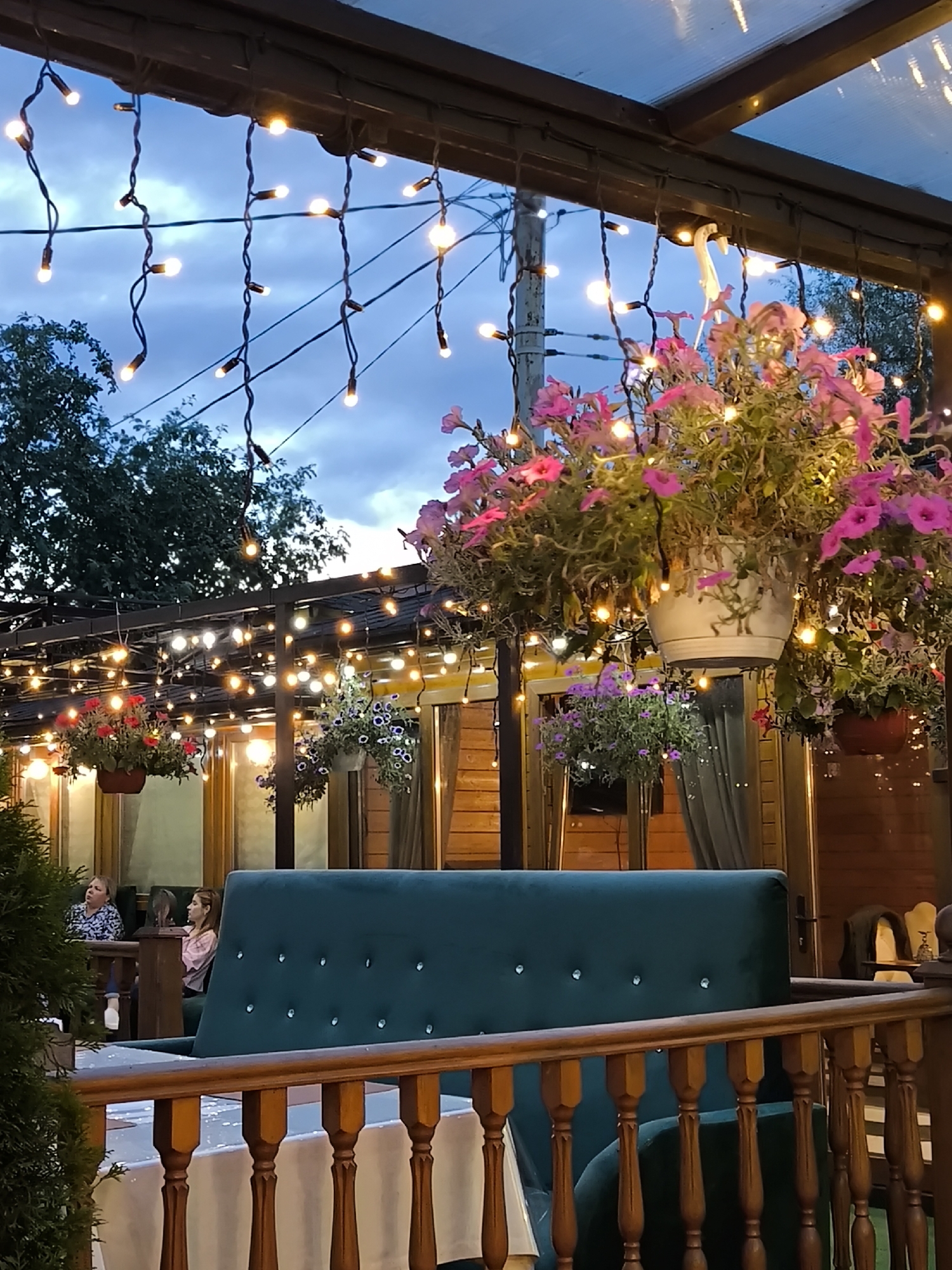 The image captures an outdoor seating area of a restaurant or cafe, likely in Khimki, Russia, at dusk. String lights are draped across the canopy and along the perimeter of the outdoor space, casting a warm glow against the darkening twilight sky. Several potted plants with vibrant pink and purple flowers hang from the ceiling, adding a touch of color and life to the scene.
In the foreground, a dark green velvet sofa with diamond-like embellishments is partially visible, along with a wooden railing and a table covered with a white tablecloth. In the background, two women are seated at tables, one looking up and away from the camera, while the other is looking down. The wooden structure of the building, visible behind the seating area, suggests a rustic or traditional architectural style. The overall atmosphere is cozy and inviting, suitable for an evening gathering. The weather appears to be calm, with a cloudy sky indicating the transition from day to night. There is no visible text in the image.