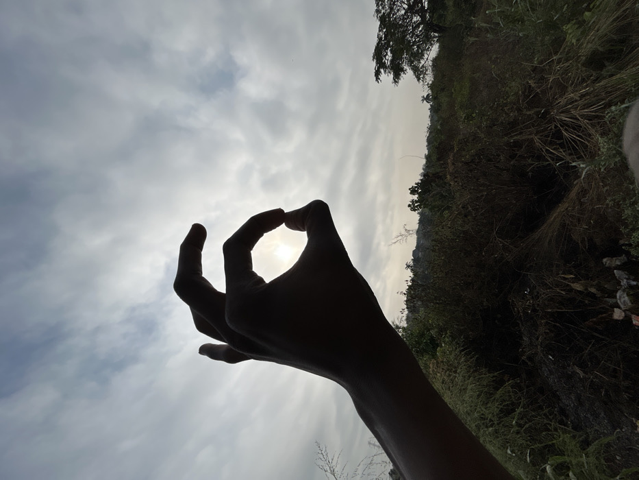 A single human hand, silhouetted against a bright sky, occupies the left foreground. The thumb and index finger are connected to form an approximately circular opening, through which a brilliant, circular light source (the sun) is centrally visible. The remaining fingers are flexed upwards. The midground consists of dense, overgrown vegetation and tall dry grasses, with discernible refuse material present at the lower left. In the background, a horizon line reveals more distant foliage and trees, including a prominent large tree on the far right. The sky is largely covered by thin, scattered clouds. This scene is situated in Debasco, Nigeria.