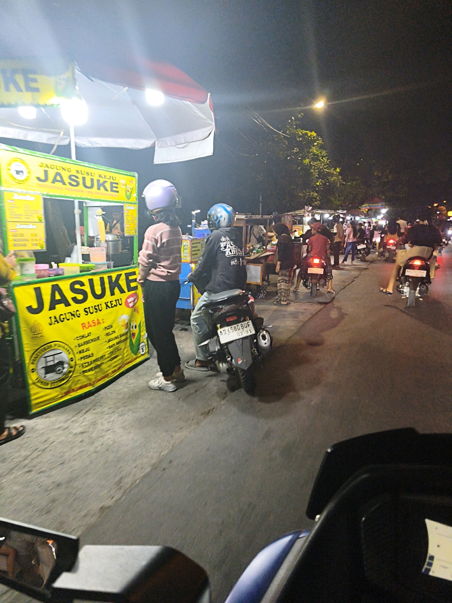 The image depicts a busy night scene in Surakarta, Indonesia, likely a street market or food stall area. In the foreground, a yellow banner advertises "JASUKE" (Jagung Susu Keju - Corn Milk Cheese), a popular Indonesian snack. Two individuals are visible at the stall, one wearing a purple helmet and a striped sweater, the other a blue helmet and a black t-shirt. They appear to be customers. Behind them, several people on motorcycles are waiting in line or passing by. The atmosphere is lively, with food stalls and streetlights illuminating the scene. The overall impression is one of a vibrant and bustling night market in Surakarta.