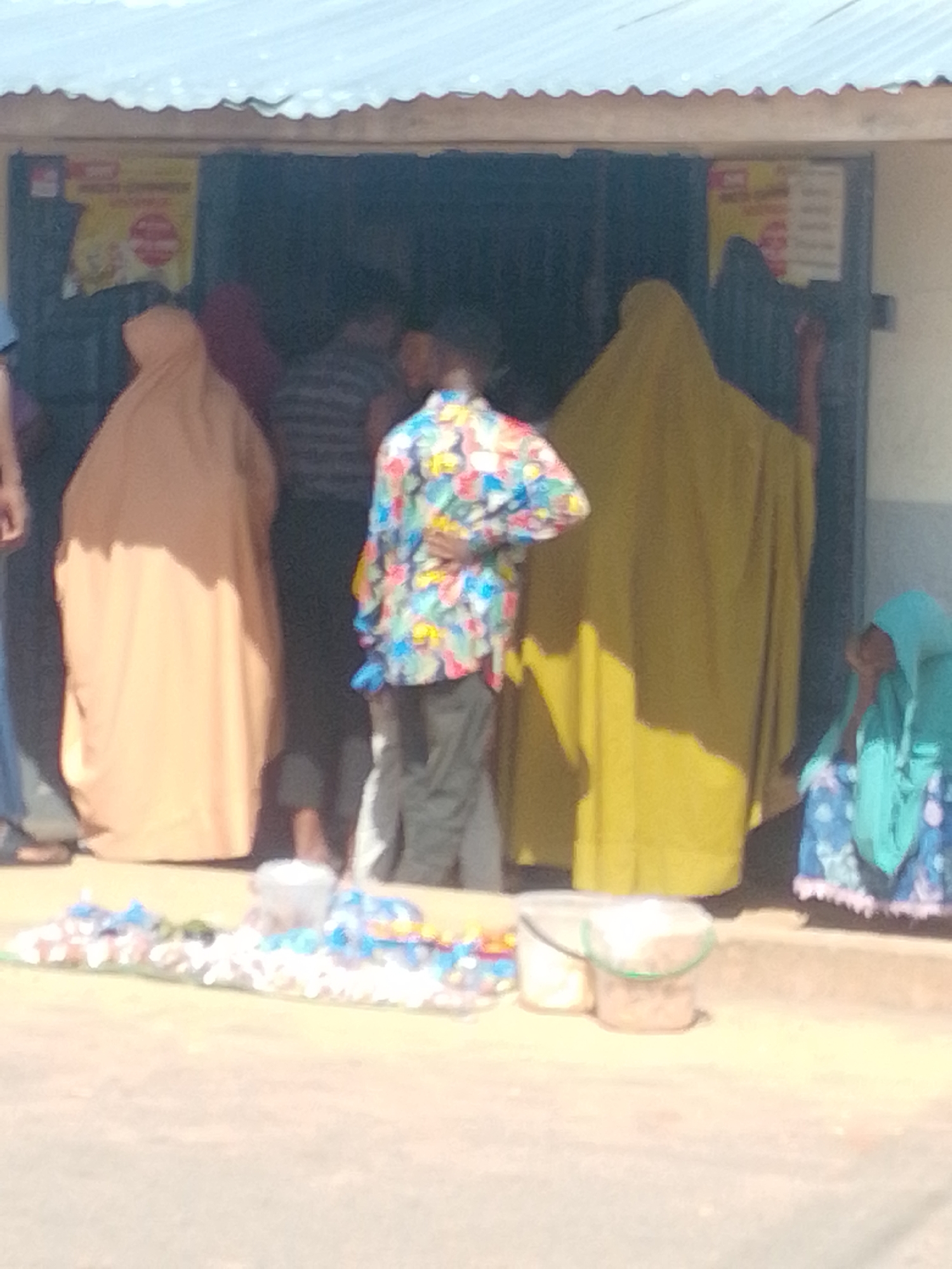 The image depicts a street scene outside a shop in Tempure, Nigeria. Several people are gathered, some appearing to be customers and others possibly shopkeepers. A person wearing a colorful, patterned shirt is standing in the foreground, facing away from the camera. To their left, two individuals are partially obscured, one in a tan hijab and another in a dark garment. To the right, a person in a bright yellow hijab stands near the shop entrance. Further to the right, a child is seated, wearing a turquoise hijab and a patterned blue and white dress. In front of the shop, a collection of items, possibly snacks or small goods, are laid out on the ground, along with two buckets. The shop itself appears to have a corrugated metal roof and an open doorway. The ground is a light-colored, sandy or dusty surface. The overall lighting suggests it is daytime, with shadows indicating sunlight.
