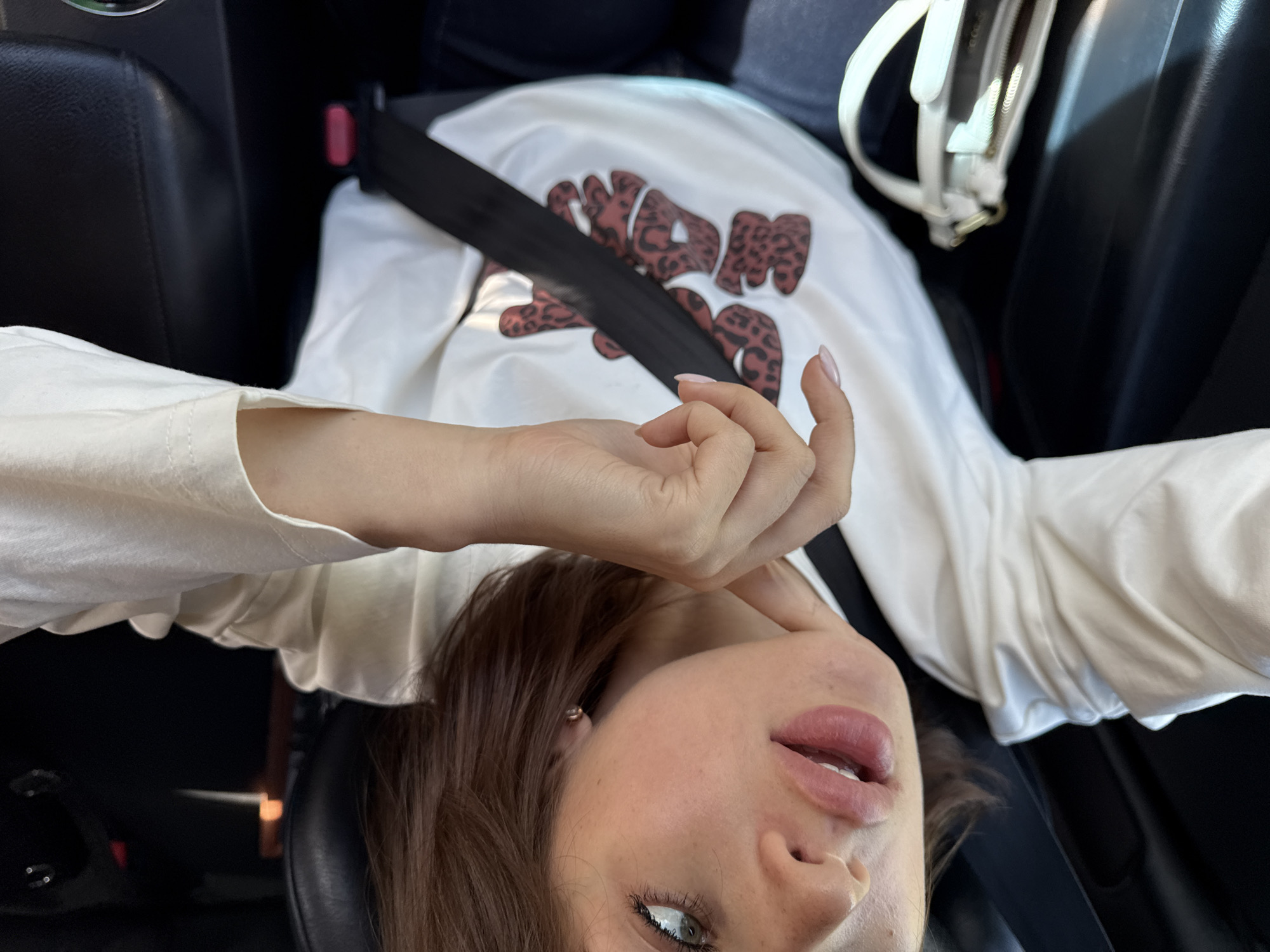 A Caucasian woman is shown from a low angle, likely in a car. She is wearing a white long-sleeved shirt with the letters "WANAM" in brown leopard print. A seatbelt crosses her chest. Her right hand is raised near her face. She has brown hair and blue eyes. The scene appears to be inside a car with black leather seats. The exact location in the city of Chaikovsky, Russia, cannot be determined from this image alone.