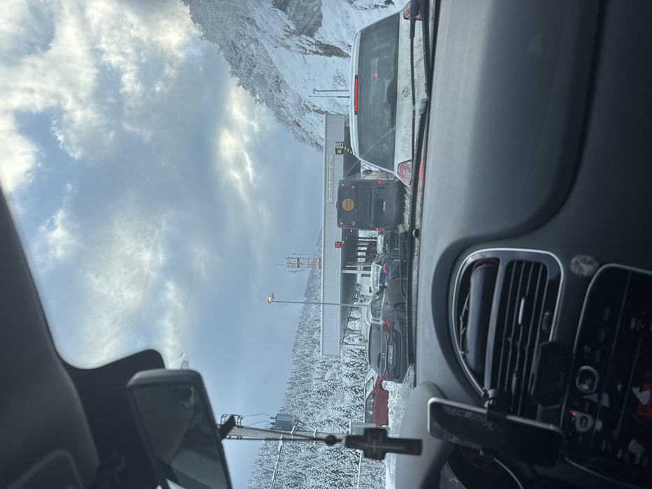 The image depicts a vehicle's interior viewpoint looking forward at the Špiljani-Mehov Krš border crossing, situated between Tutin Municipality, Serbia, and Rožaje Municipality, Montenegro. The environment is a snow-covered mountainous region, with coniferous trees heavily laden with snow lining the hillsides.
In the foreground, the grey dashboard of the observer's vehicle is visible, including air vents, portions of the steering wheel, and a mounted smartphone on the left. A small crucifix ornament hangs from the interior rearview mirror support.
The primary subject is the multi-lane border crossing facility, characterized by a broad, light-colored canopy extending over several inspection lanes. Multiple vehicles are queued. Directly ahead is a white hatchback, its rear windshield partially obscured by dirt or moisture. To its right, a black bus with a circular yellow emblem on its rear is visible. To the left of the white hatchback, a dark grey Mazda station wagon displays a Bosnian license plate "BA-00710". Red 'X' symbols are prominently displayed above some of the border lanes, indicating they are closed.
In the background, a red and white telecommunications tower is visible on a snow-covered hill to the left of the crossing structure. A single street light pole stands near the tower. The sky is overcast with varied cloud formations, showing patches of brighter light through the grey clouds. No individuals are clearly visible, but the scene implies human presence within the queued vehicles.