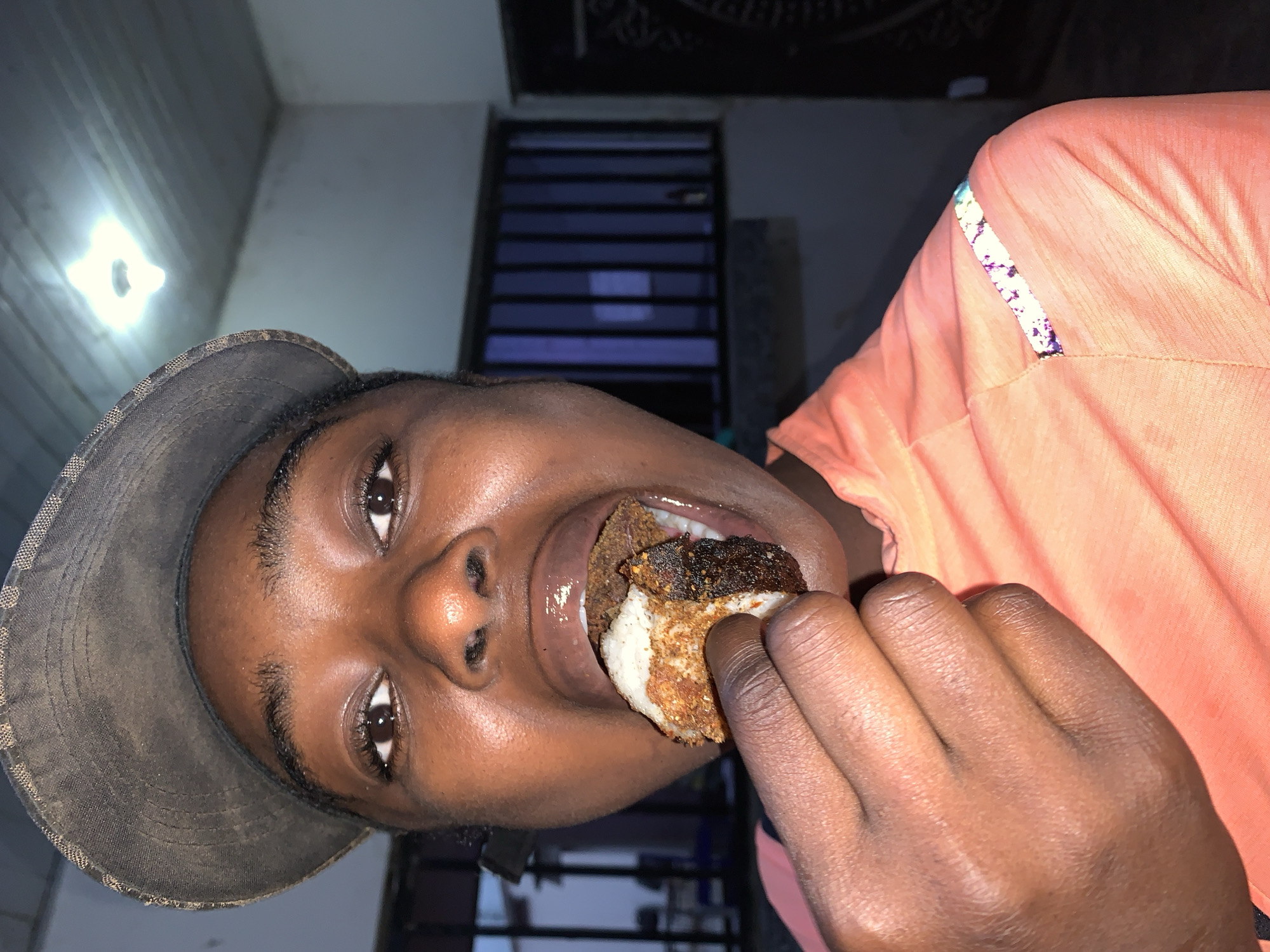 A person is shown at a close, angled perspective, eating what appears to be a round food item with a light bottom and dark top layer. The person is wearing a brown hat and an orange shirt. The background shows what appears to be an interior location, with a light fixture and metal bars. The context of New Stadium, Nigeria is external information, and therefore not included in this description.