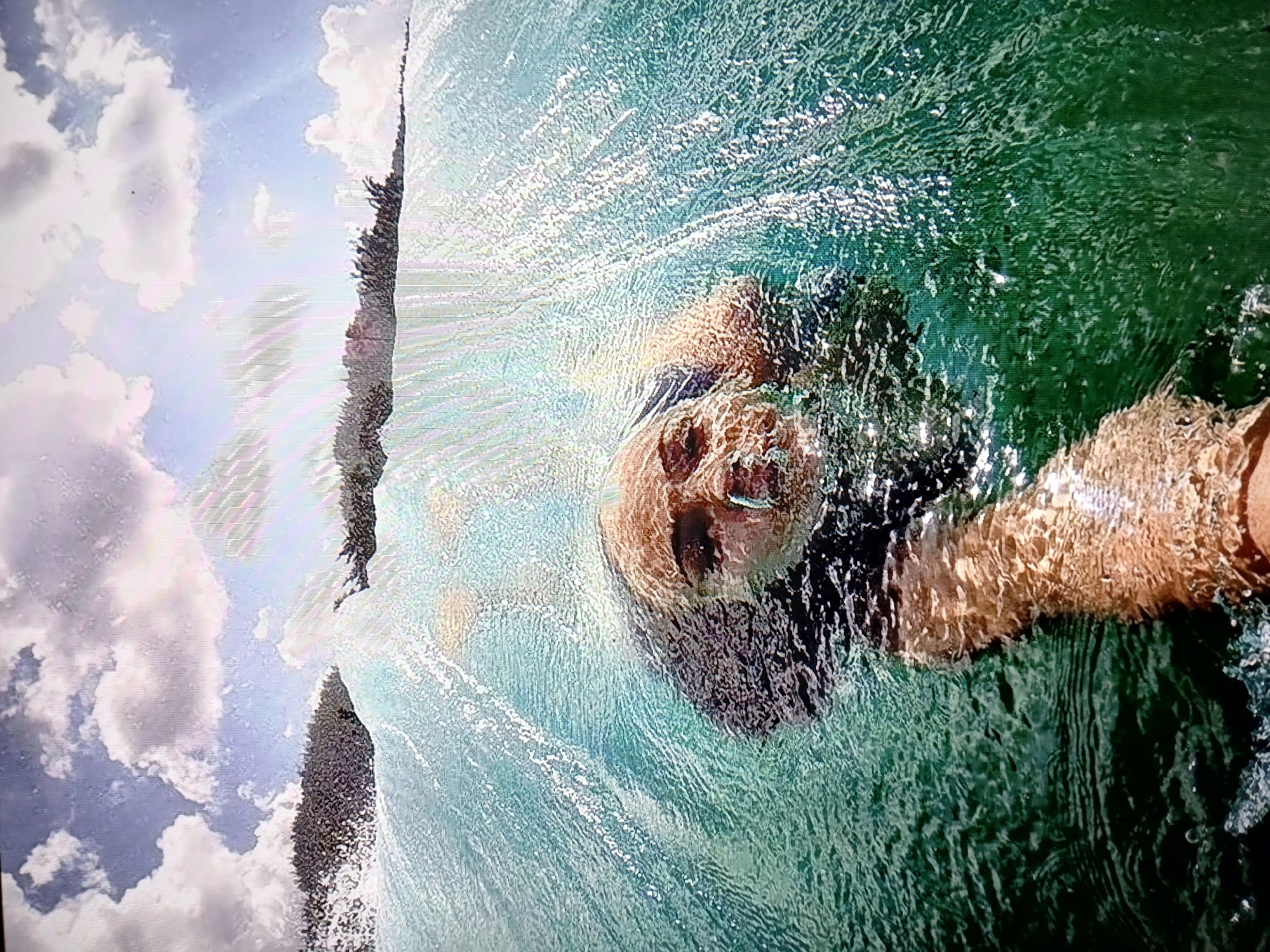 In this underwater selfie, a person is partially submerged in a body of water, with their face closest to the camera. The person's eyes are open, and they appear to be looking at the camera. Bubbles are visible around their face, suggesting they are holding their breath or exhaling underwater. Their arm is extended forward, holding the camera. The water is a clear turquoise color, with sunlight creating bright highlights and reflections on the surface. Behind the person, the water surface curves upwards, revealing a glimpse of the sky with scattered white clouds and the sun's rays breaking through. The background also shows a blurred line of vegetation, possibly trees or bushes, along the horizon. The overall impression is an action shot of someone enjoying a day in the water, possibly swimming or surfing.