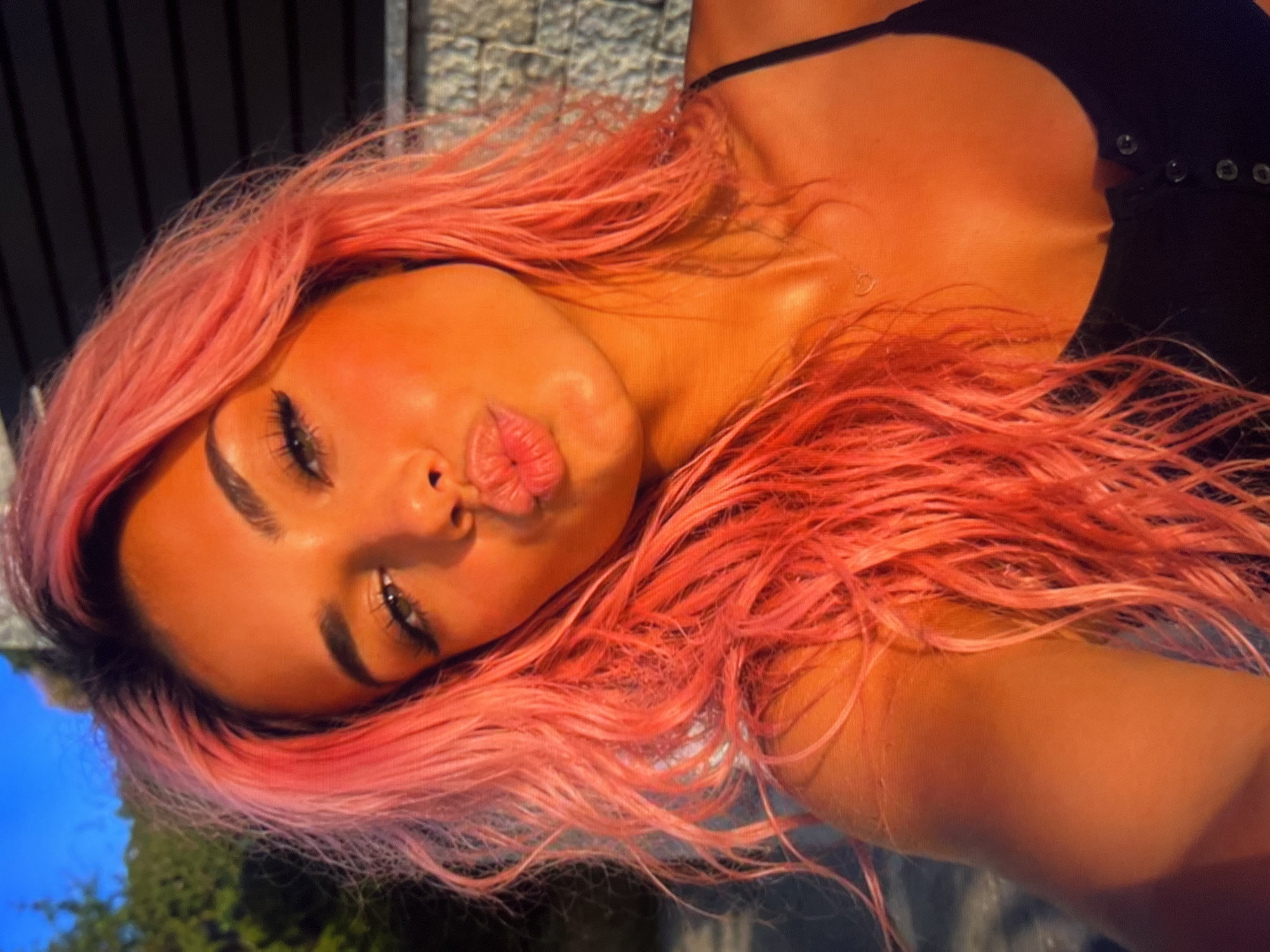 A close-up, shoulder-up, selfie-style photograph shows a young woman with vibrant pink hair and striking makeup. Her lips are pursed in a kiss, and her eyes are framed by dramatic eyeliner and long lashes. She wears a black tank top, and a delicate necklace is visible on her collarbone. The lighting suggests it is sunset, casting a warm, orange glow over her skin and hair, and highlighting the texture of the brick wall behind her. Trees and a sliver of blue sky are visible in the upper left corner.