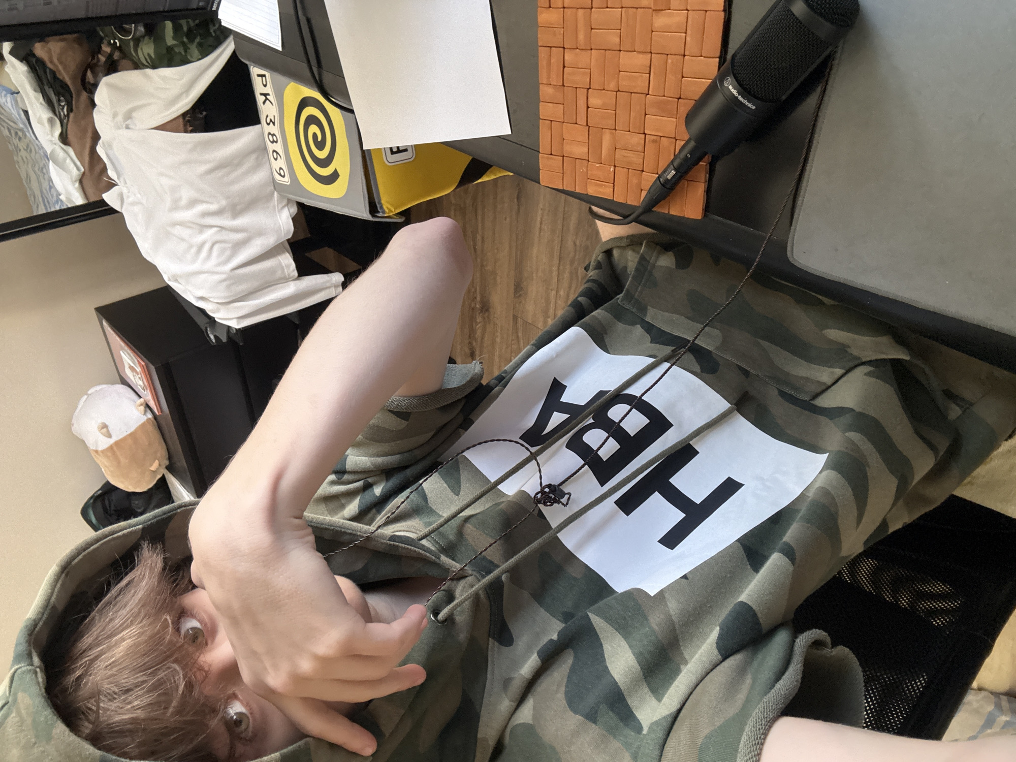 A young person with brown hair, wearing a camouflage hoodie with "HBA" printed in large white letters, is shown from the chest up. Their right hand is raised, partially covering their mouth. The background is cluttered, with a dresser on the left, a white t-shirt draped over a chair or desk in the middle, and a yellow box with a stylized spiral logo on the right. A black microphone with "Audio-Technica" written on it is visible on a desk in the foreground, next to an orange brick-patterned surface and a gray mousepad. The environment appears to be an indoor space, possibly a bedroom or office. There is no indication of the location being in Khimki, Russia.