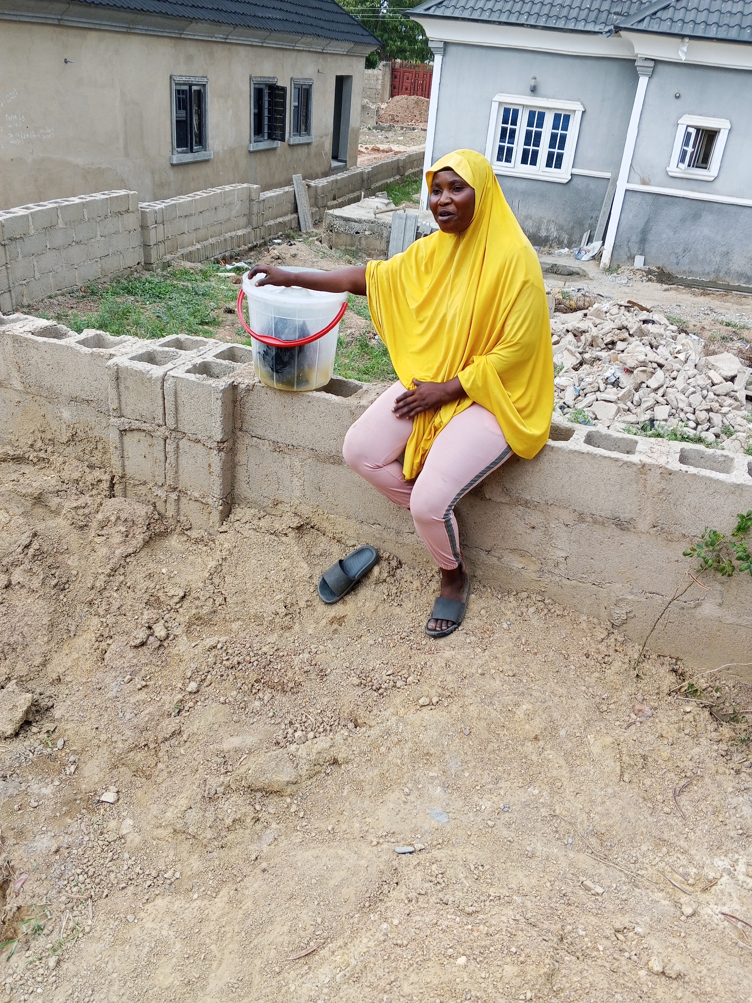 This image captures a woman seated on a low concrete block wall in what appears to be a developing or construction area in Adonkolo, Nigeria, under natural daylight conditions.
The main subject is a woman with dark skin, wearing a bright yellow hijab that covers her head and shoulders, and light pink athletic-style pants with dark stripes down the sides. She is also wearing dark sandals on her feet, with one slipper discarded on the ground nearby. Her expression is neutral as she looks towards the right side of the frame. She is holding a transparent plastic bucket with a red handle in her left hand, which contains some dark items.
She is sitting on a section of a concrete block wall, which extends across the midground. In front of the wall, the ground is predominantly rough, light brown soil or sand, with some scattered pebbles and sparse patches of green grass. Behind her, the ground is similarly uneven, with piles of construction debris, including broken concrete and rubble, visible to her right.
In the background, there are two houses in different stages of completion. To the left, a light brown, possibly unpainted or rendered house with a dark roof and multiple dark-framed windows with louvers. Faint white chalk-like writings, possibly construction notes or measurements, are visible on its wall. To the right, a grey-painted house with a dark roof and white-framed windows, appearing more finished. The overall setting suggests an urban or semi-urban area undergoing development or expansion. The weather appears fair, with no harsh shadows indicating a bright or slightly overcast day.