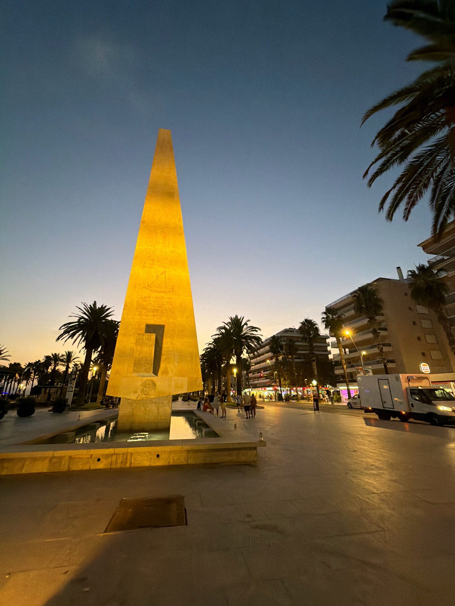 This is a photograph of the Monument to Jaime I in Salou, Spain, captured at dusk. The monument, a large, golden obelisk with an angular, pyramidal shape, stands prominently in the foreground. It features a cutout in the shape of the number "1" and carved reliefs on its surface. The monument is situated next to a shallow water feature or fountain, with reflections visible on the wet surfaces.
To the right of the monument, a paved pedestrian promenade stretches into the background, lined with palm trees and modern buildings. The buildings appear to be apartments or hotels, with balconies and illuminated windows. Streetlights and lights from shops cast a warm glow on the promenade, illuminating the area. A white van and several parked cars are visible on the right side of the promenade.
Several individuals are present on the promenade, walking and standing. They are casually dressed, suggesting a leisurely evening atmosphere. The sky above is a gradient of deep blue and fading orange, indicative of twilight. The overall scene depicts a lively public space in a coastal town at the end of the day.