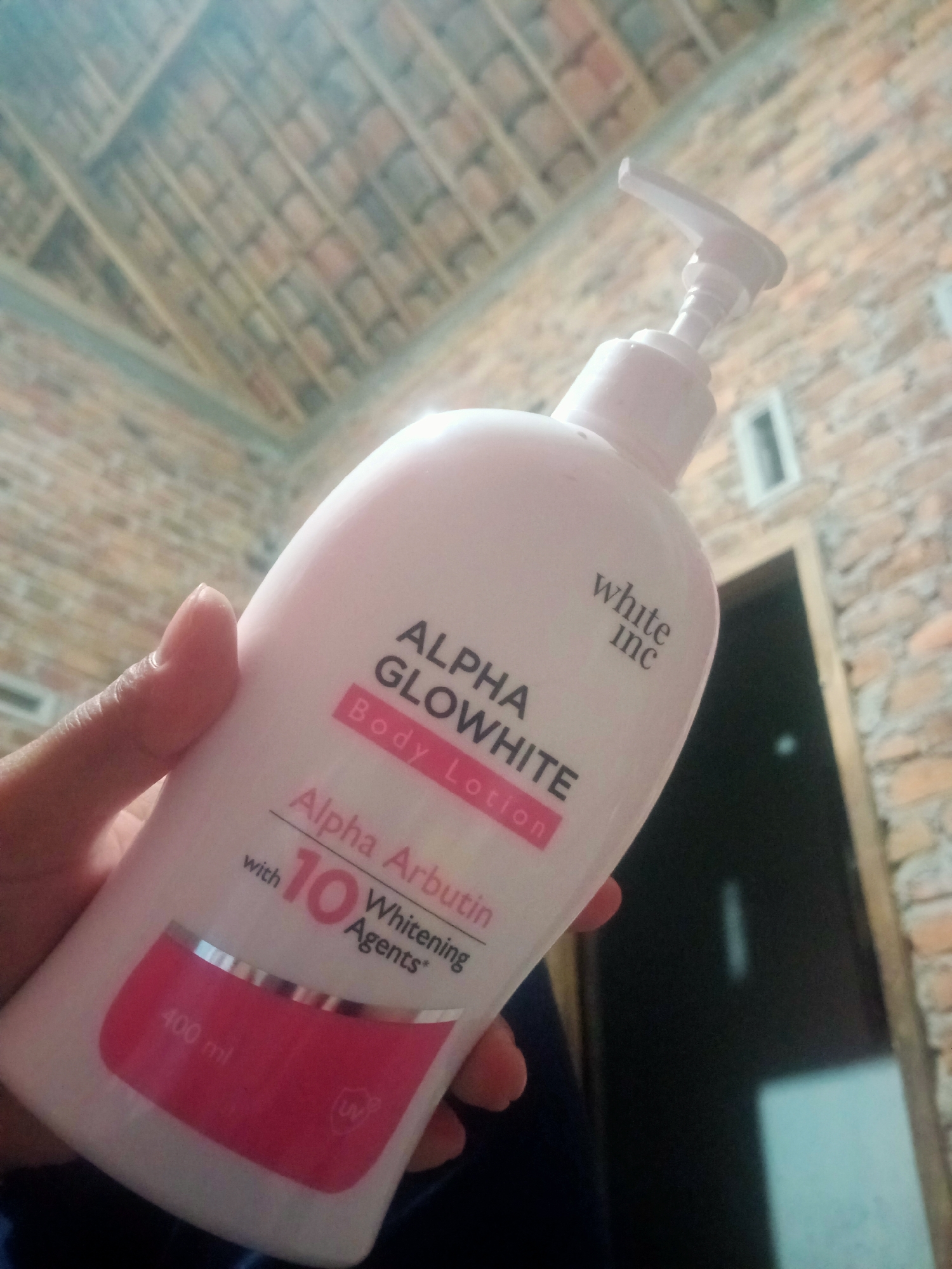 A close-up shot shows a person's hand holding a pink and white bottle of "ALPHA GLOWWHITE BODY LOTION" with "Alpha Arbutin" and "10 Whitening Agents" printed on it. The bottle has a white pump dispenser and a silver band around the bottom that says "400 ml". A "UV" symbol is visible on the bottle. The background consists of a brick wall and a ceiling with exposed wooden beams. The image appears to have been taken indoors in Nusa Agung, Indonesia.
