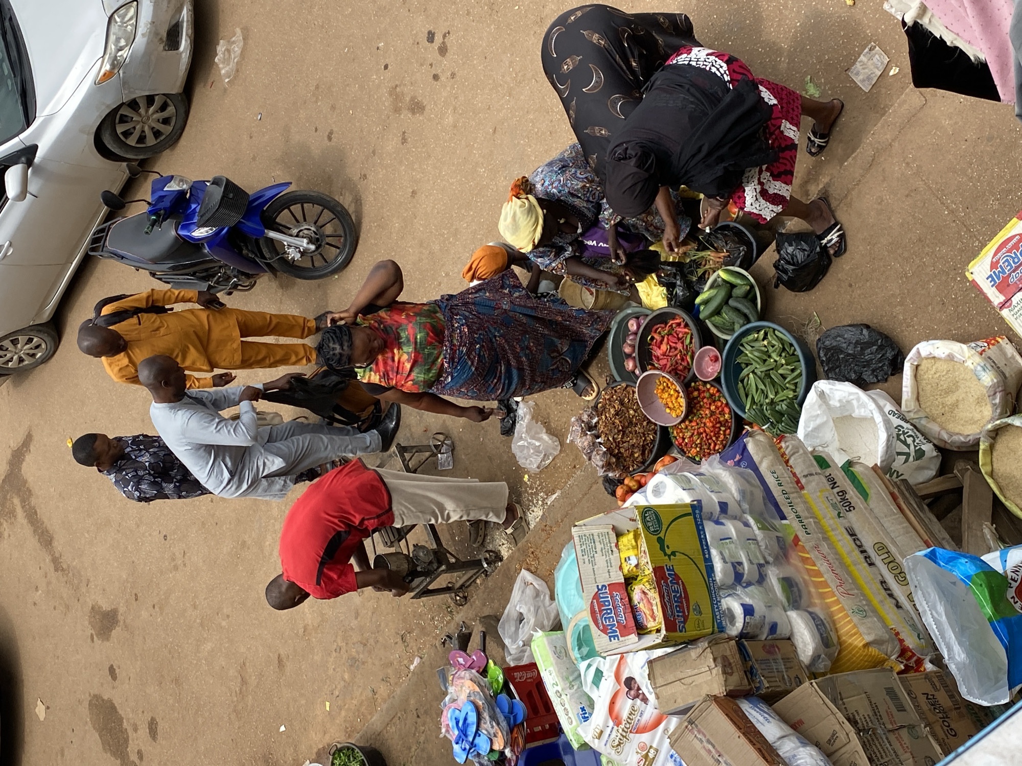 Captured from an elevated viewpoint, the image depicts a marketplace scene in Bukuru, Nigeria. Various individuals are observed engaging in commerce, their activities clustered around a collection of goods and vehicles.
Several people are situated near an array of fresh produce, including tomatoes, peppers, cucumbers, and okra, displayed in basins. Two women, one wearing a patterned skirt and top and the other a black garment with crescent moon motifs, are focused on the produce. Nearby, a woman lies on her side.
In proximity to the group, a man in a red shirt is seated, while another man in a gray shirt is standing. Further away, a man in a yellow shirt is also seen.
Adjacent to the group, there is a car and a blue motorcycle. To the side, stacked containers of various products, including boxes labeled "Supreme" and "Softcare," are visible, along with bags of rice labeled "MR. RICE." A Coca-Cola crate also appears in the scene.