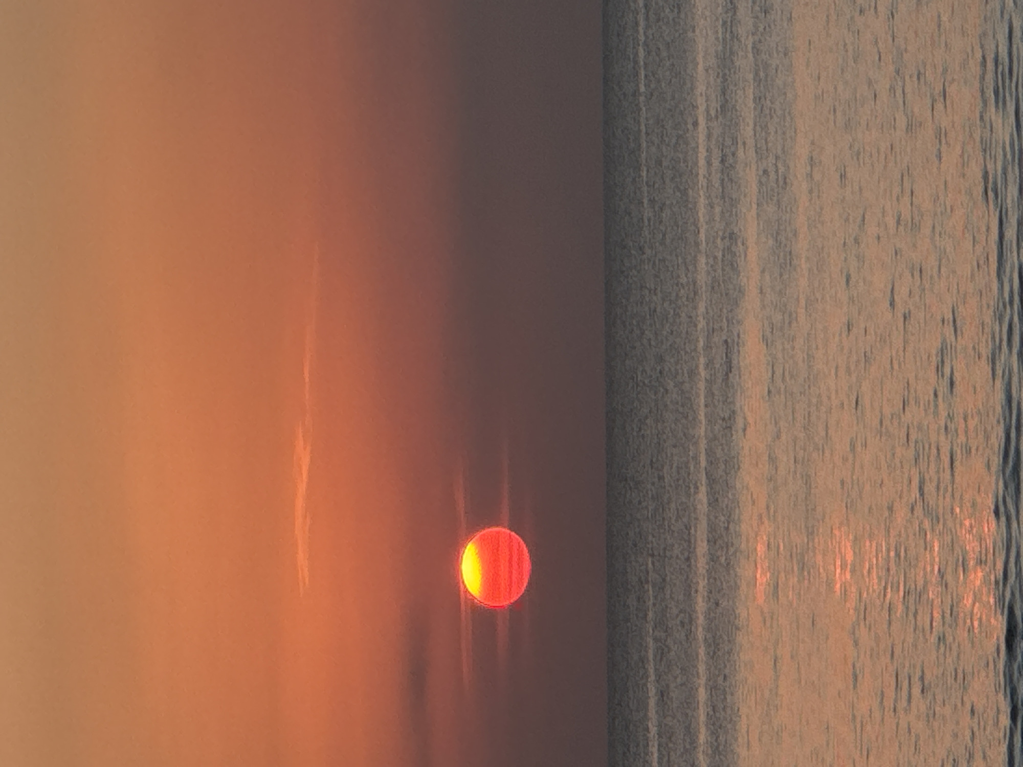 The image depicts a sunset over the ocean in Kuta Selatan, Indonesia. The sun, a vibrant orb of red and yellow, is partially obscured by thin clouds and hovers just above the horizon. The sky transitions from a warm orange at the top to a muted purple-grey closer to the water. The ocean is calm, with gentle ripples reflecting the colors of the sunset, creating a shimmering path of light from the sun to the foreground. The scene is natural and serene, with no human presence or man-made objects visible.