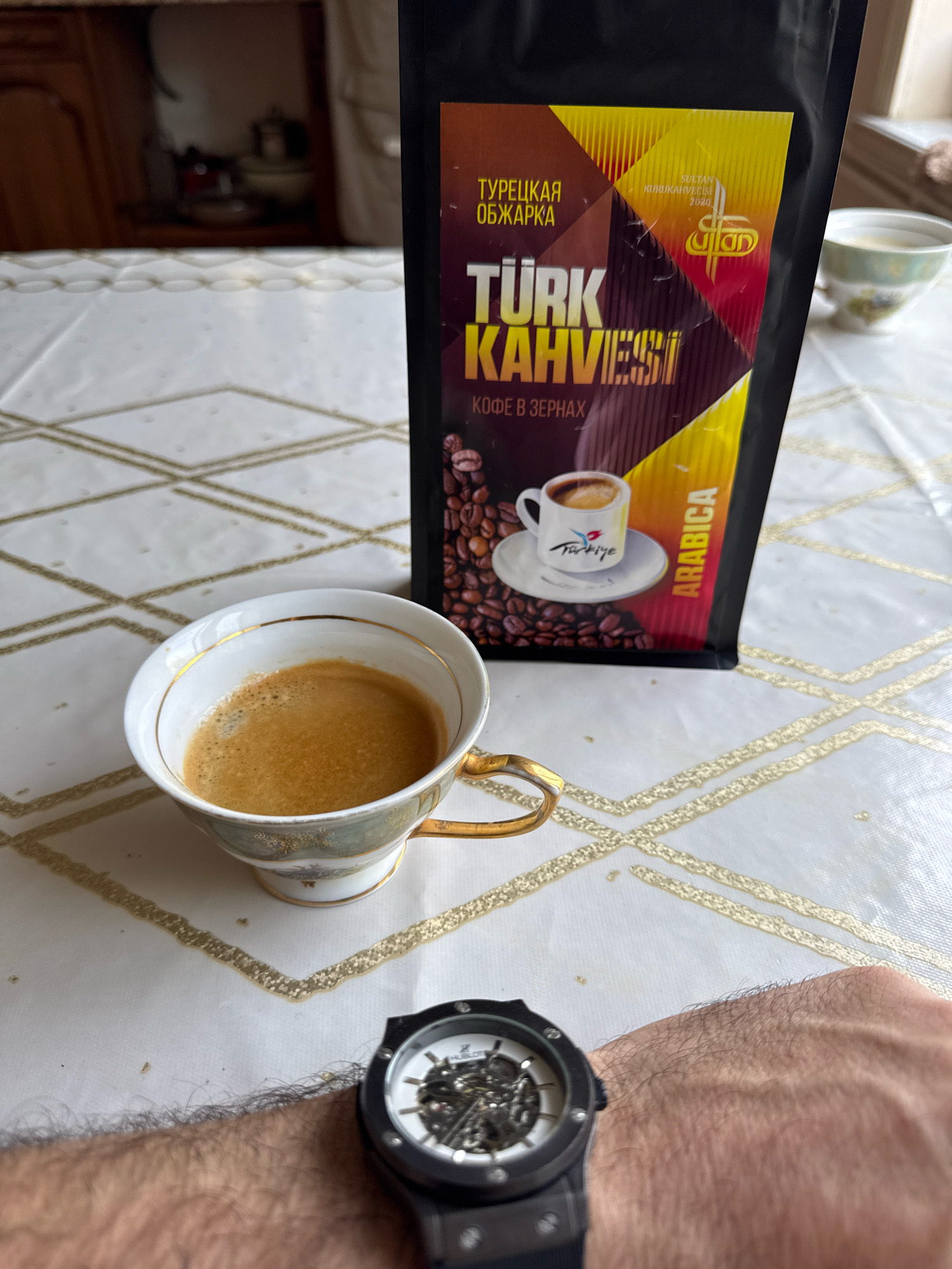 The image shows a close-up view of a table setting with a cup of Turkish coffee, a bag of Turkish coffee beans, and a watch on a person's wrist. The coffee is in a decorative cup with gold trim and filled with a frothy dark liquid. The coffee bag is black with colorful text and graphics, indicating "Türk Kahvesi" and "Arabica." A small white saucer with a miniature cup of coffee is depicted on the bag. The table is covered with a white tablecloth featuring a gold diamond pattern. In the background, a blurred cabinet is visible. The watch on the wrist has a black band and a skeletonized face, revealing intricate inner workings. The overall scene suggests a moment of relaxation and enjoyment of coffee. The image does not contain any visual information that would place it in the city of Belidzhi, Russia.