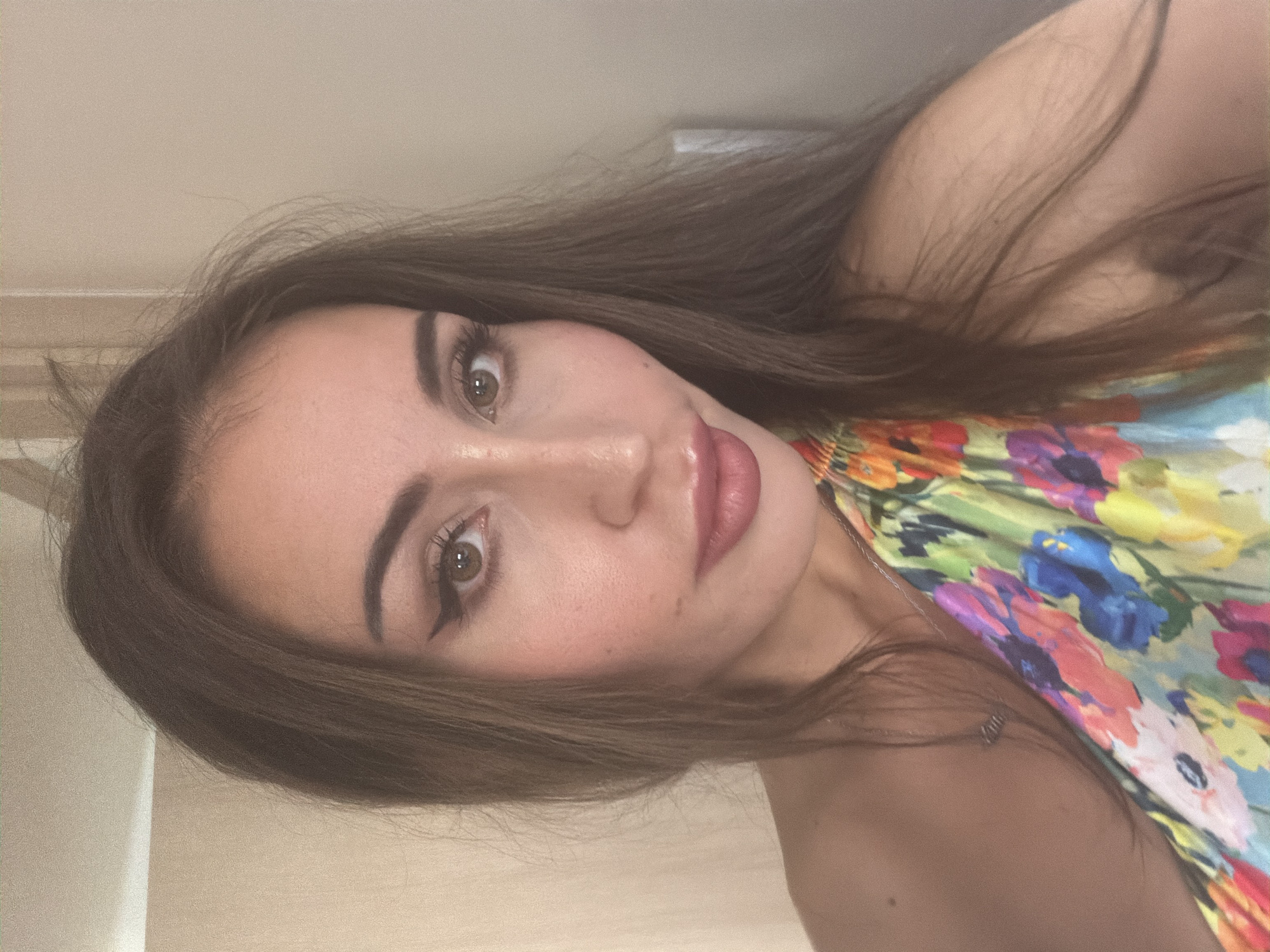 A woman with long brown hair and brown eyes poses for a selfie. She is wearing a brightly colored floral top that is off-the-shoulder. Her makeup includes winged eyeliner and pink lipstick. A silver necklace is visible around her neck. The background appears to be an indoor setting with a neutral-colored wall. The image was taken in Tersefanou, Cyprus.