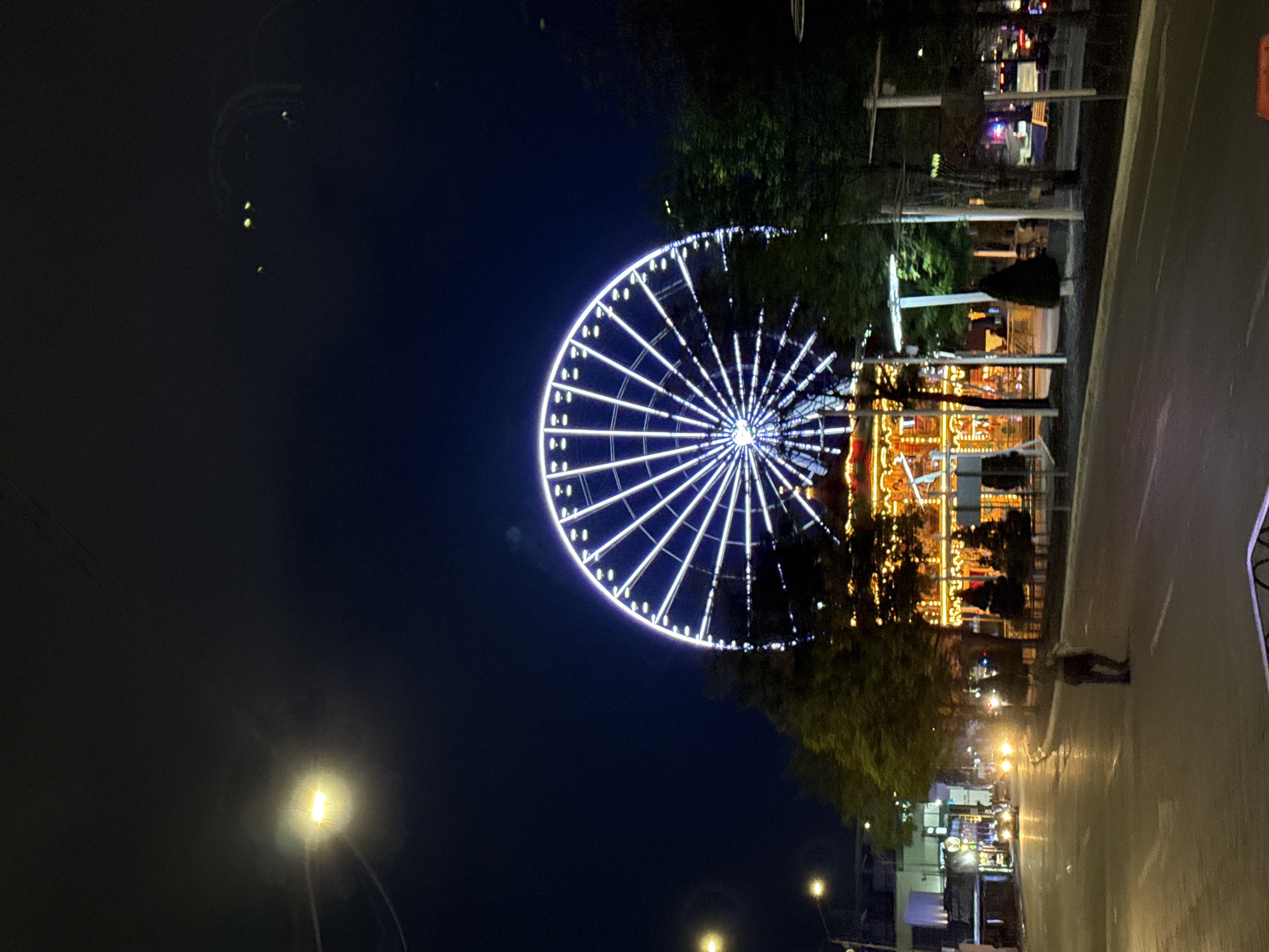The image shows a nighttime view of a ferris wheel illuminated with white lights, located in Puebla, Mexico. The wheel is on the left side of the frame against a dark blue sky. To the right of the wheel, there's a lit carousel and other amusement park structures, partially obscured by trees. A person walks on the sidewalk to the right of the ferris wheel. Streetlights are visible along the road, and buildings and other structures are visible in the background.