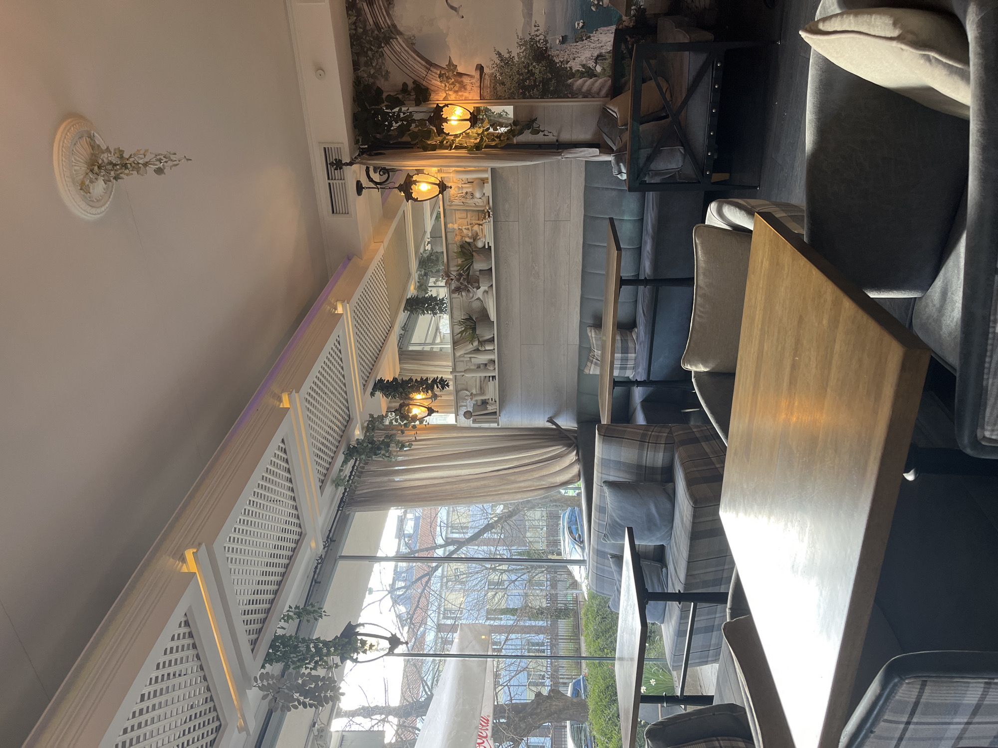The interior of a restaurant in Burgas, Bulgaria is shown. The perspective is from within the restaurant, looking outwards through a large window. Outside, there is a view of buildings and trees, suggesting an urban setting. Inside, there are tables and chairs arranged for dining. There are no people visible in the image. The restaurant has a sophisticated decor, with ornate light fixtures and decorative elements.