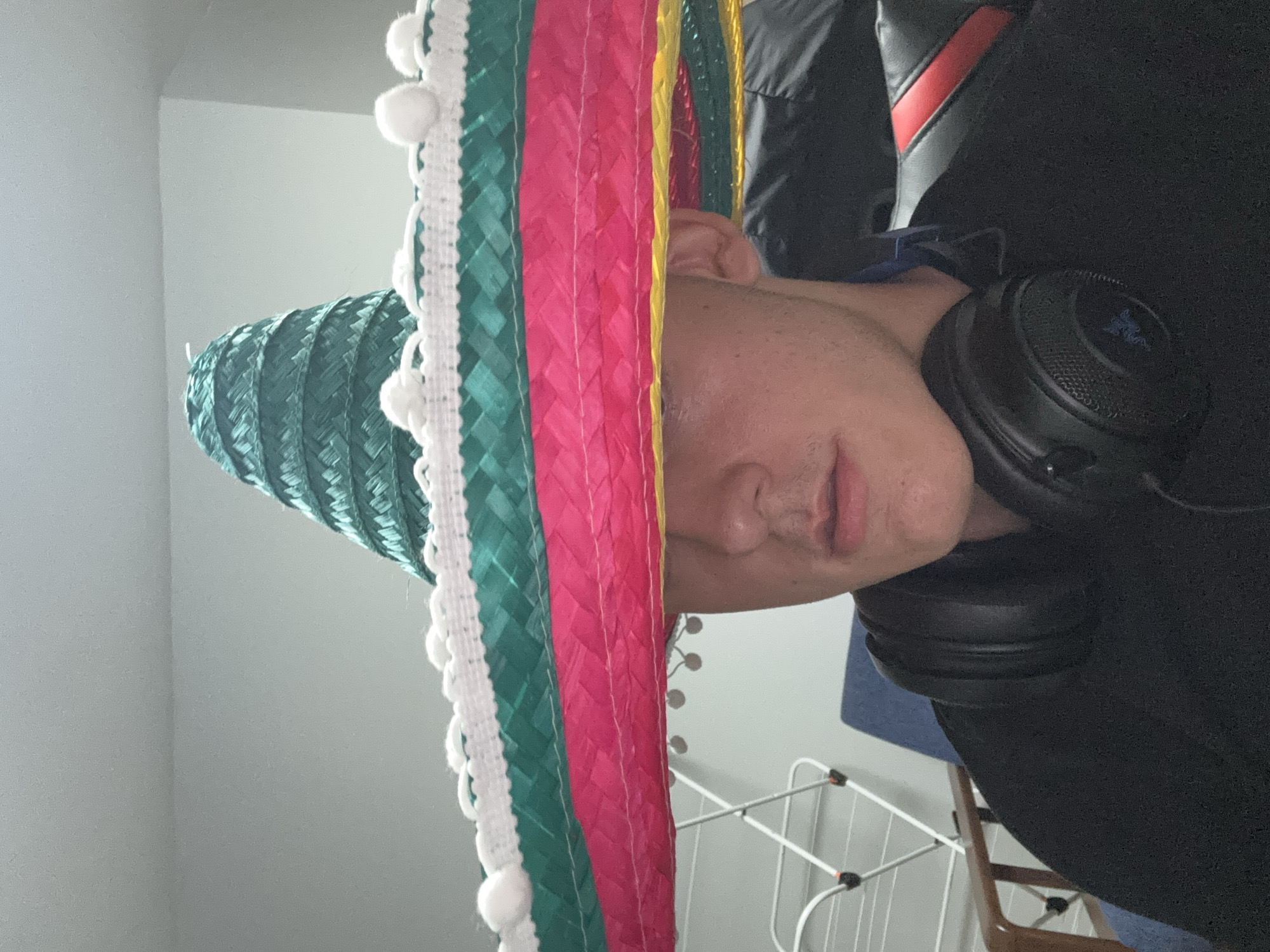 The image depicts a person wearing a sombrero and headphones. The sombrero is multi-colored with white pom-poms. The person is wearing a black shirt. The background includes a wall and a drying rack. There is no indication of the location being in Plovdiv, Bulgaria.