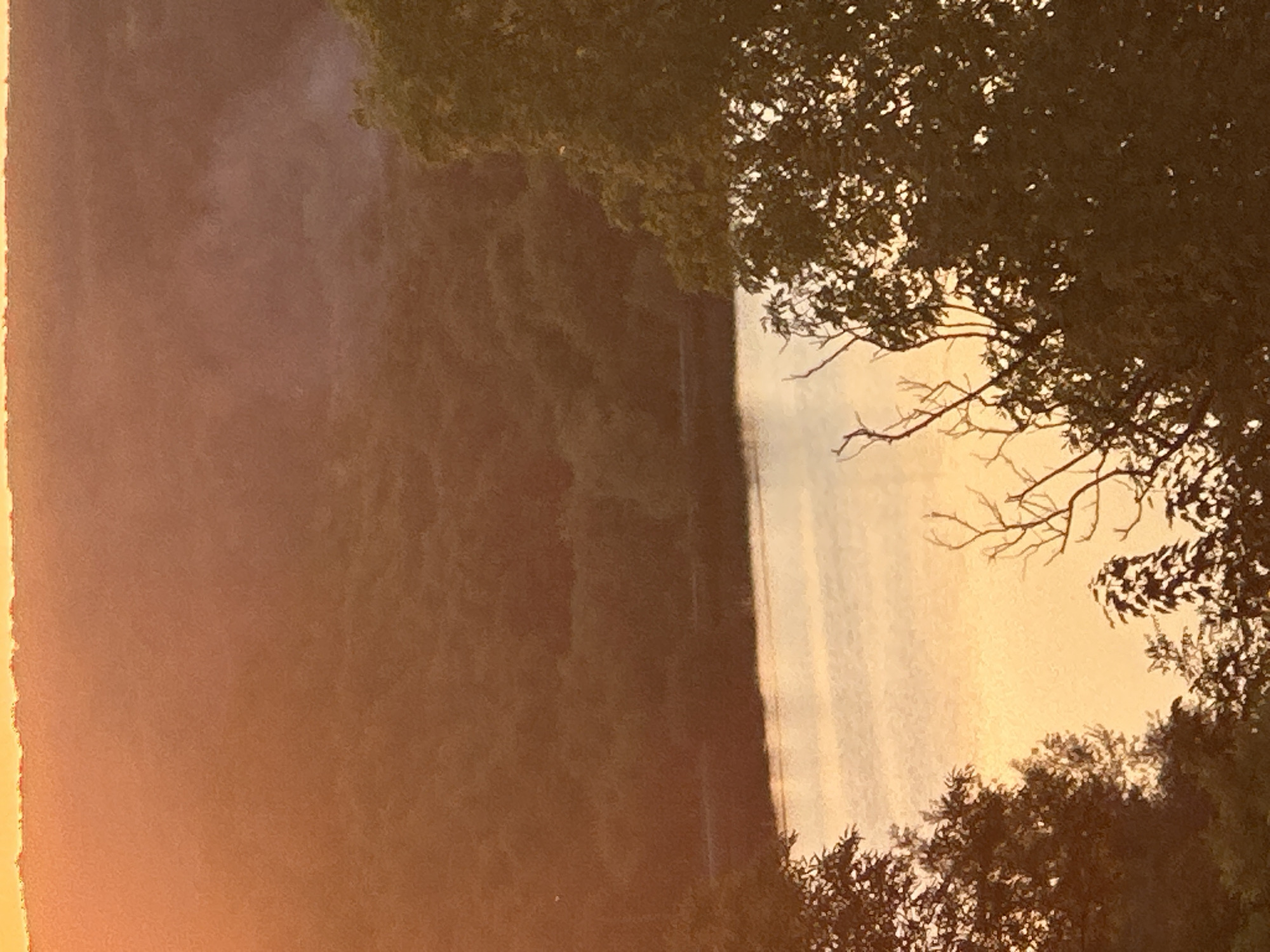A high-angle, medium shot captures a serene landscape at sunset, likely on the outskirts of Makiv, Ukraine. The scene is dominated by a body of water, possibly a river or lake, reflecting the warm hues of the setting sun. Lush, dark green foliage frames the water from the left and right foreground, with a distant, undulating treeline receding into a hazy, orange-tinted sky. A thin wisp of smoke rises from the middle ground, suggesting a small fire or industrial activity. The overall atmosphere is tranquil, highlighting the natural beauty of the area.