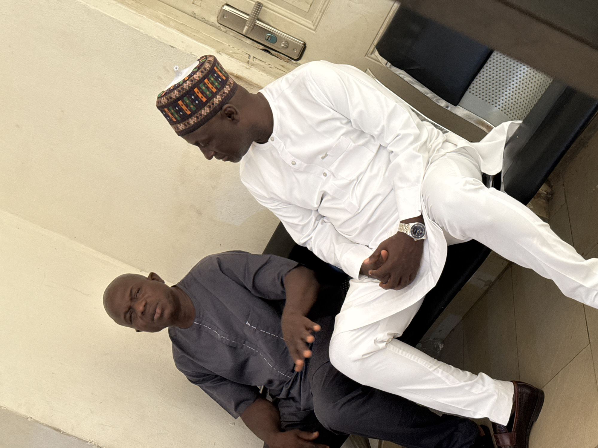 The image features two adult men seated indoors, likely in an office or waiting area in Abuja, Nigeria, given the traditional attire. The perspective is tilted, showing the scene from a high angle.
The man in the foreground, positioned on the right, wears a white long-sleeved traditional Nigerian tunic or kaftan with matching trousers. He sports a distinctive patterned cap with brown, orange, and green geometric designs. His skin tone is dark, and he wears a silver-toned wristwatch on his left wrist. He is looking downwards, with his hands clasped in front of him. His feet are clad in brown leather loafers. Behind him, a black chair with a silver metallic frame and a section with a perforated pattern is visible. A light-colored wall or door in the background features a modern silver door handle and lock mechanism.
The second man, seated on the left, wears a dark grey long-sleeved traditional tunic and dark trousers. He also has a dark complexion and appears to be looking upwards, towards the camera or slightly past it, with a neutral or slightly serious expression. His left hand rests on his lap, and his right hand is positioned on his leg.
The men appear to be either conversing or waiting together. The traditional clothing indicates a cultural context prevalent in Nigeria. The indoor setting suggests diffused light, likely artificial or ambient daylight. There is no visible text in the image.