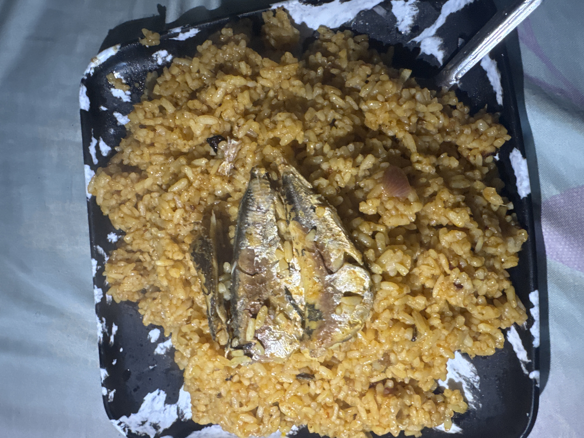 A high-angle shot captures a meal consisting of jollof rice topped with three small, whole, fried or smoked fish on a rectangular black and white splatter-patterned plate. The rice is a warm orange-brown color, indicating it has been cooked with a tomato-based sauce, and appears to be short to medium grain. Small flecks of darker material, possibly spices or vegetables, are interspersed throughout the rice. The fish, likely sardines or similar small fish, are arranged in a slight overlap towards the center of the rice pile. They exhibit a golden-brown hue with shimmering scales and some darkened areas, consistent with cooking. A metallic utensil, possibly a spoon, is visible on the bottom right corner of the plate. The plate rests on a light-colored, possibly fabric, surface with faint horizontal and vertical striped patterns, suggesting a tablecloth or bedding. There are no people visible, nor are there any discernible features in the background that would specifically identify the location as the city of Ihiagwa, Nigeria; the environment is confined to the immediate dining setting.
