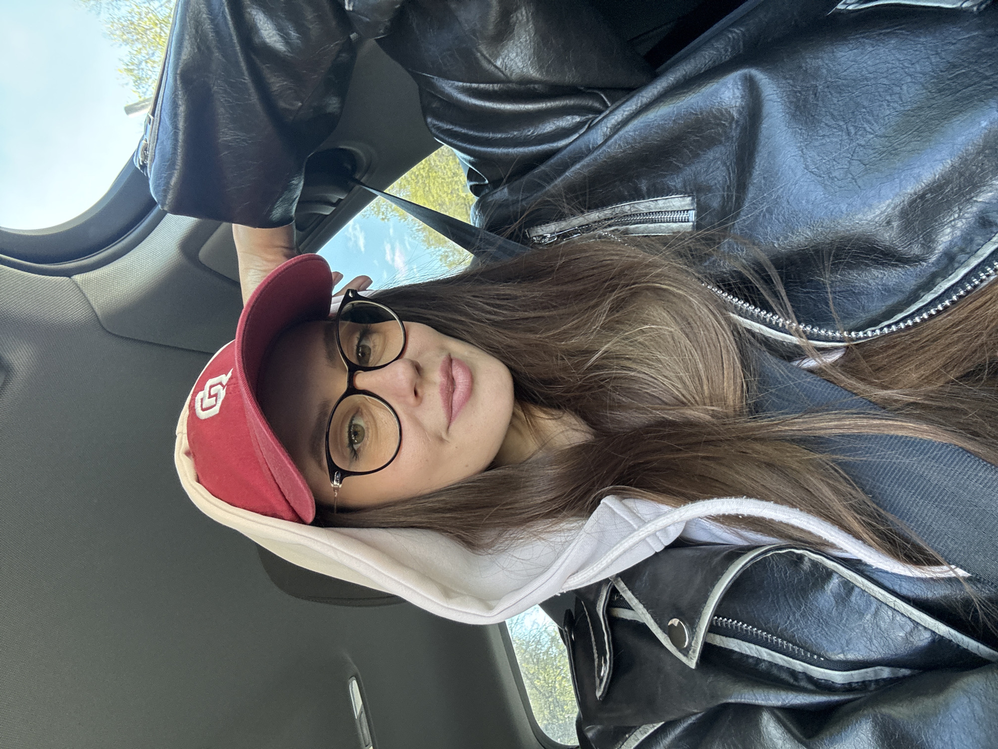 A young woman in a car, possibly the driver. She wears glasses and a red baseball cap with a white logo, a white hooded sweatshirt, and a black leather jacket. A seatbelt is visible across her torso. The background shows a car interior and a glimpse of the sky and trees through the window. Location in the city of Krasnoyarsk, Russia, cannot be determined from the provided image.