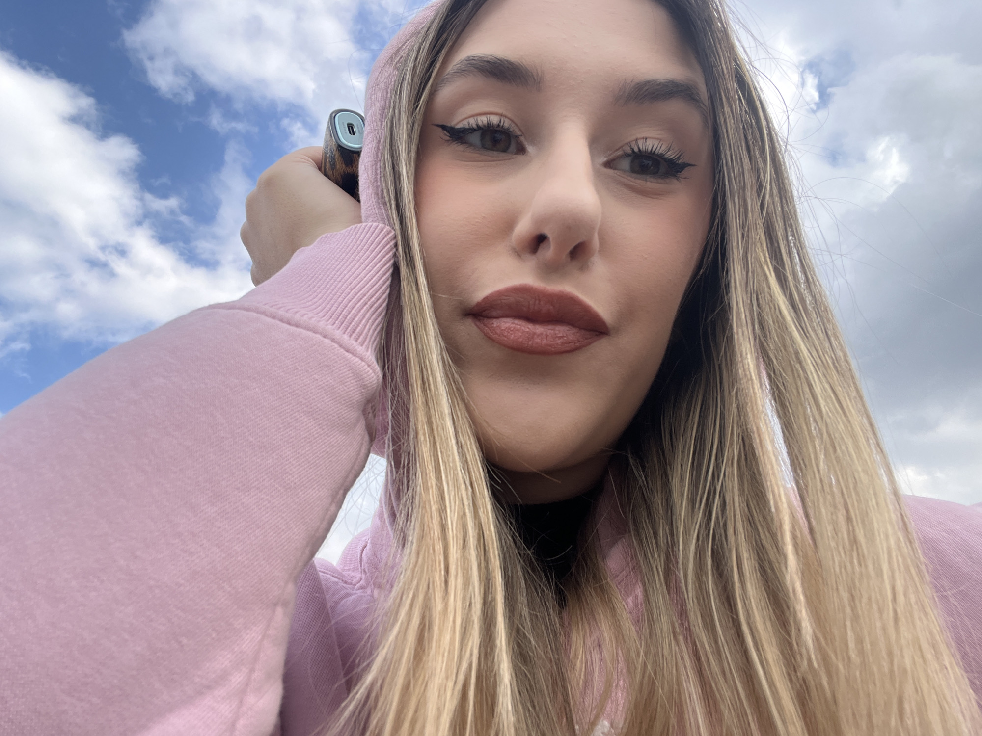 A young woman with light brown hair and wearing a pink hooded sweatshirt is holding an electronic device in her left hand. The background shows a cloudy sky. There is no information available about the location in Sofia, Bulgaria.