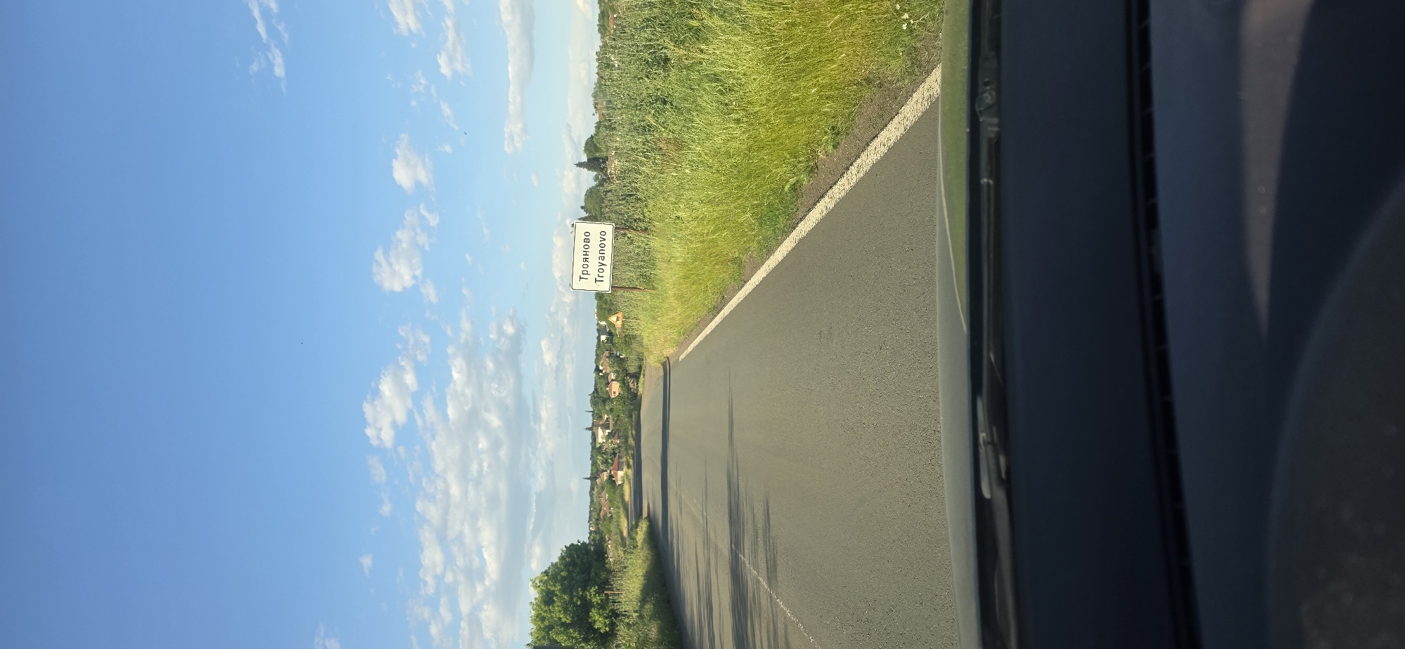 The image shows a view from inside a car driving on a road. A sign on the side of the road indicates the entrance to the city of Troyanovo, Bulgaria. The sky is blue with scattered clouds. The road is asphalt and is bordered by green vegetation on the right. The image is taken from the driver's perspective. The scene suggests a sunny day and a rural environment.