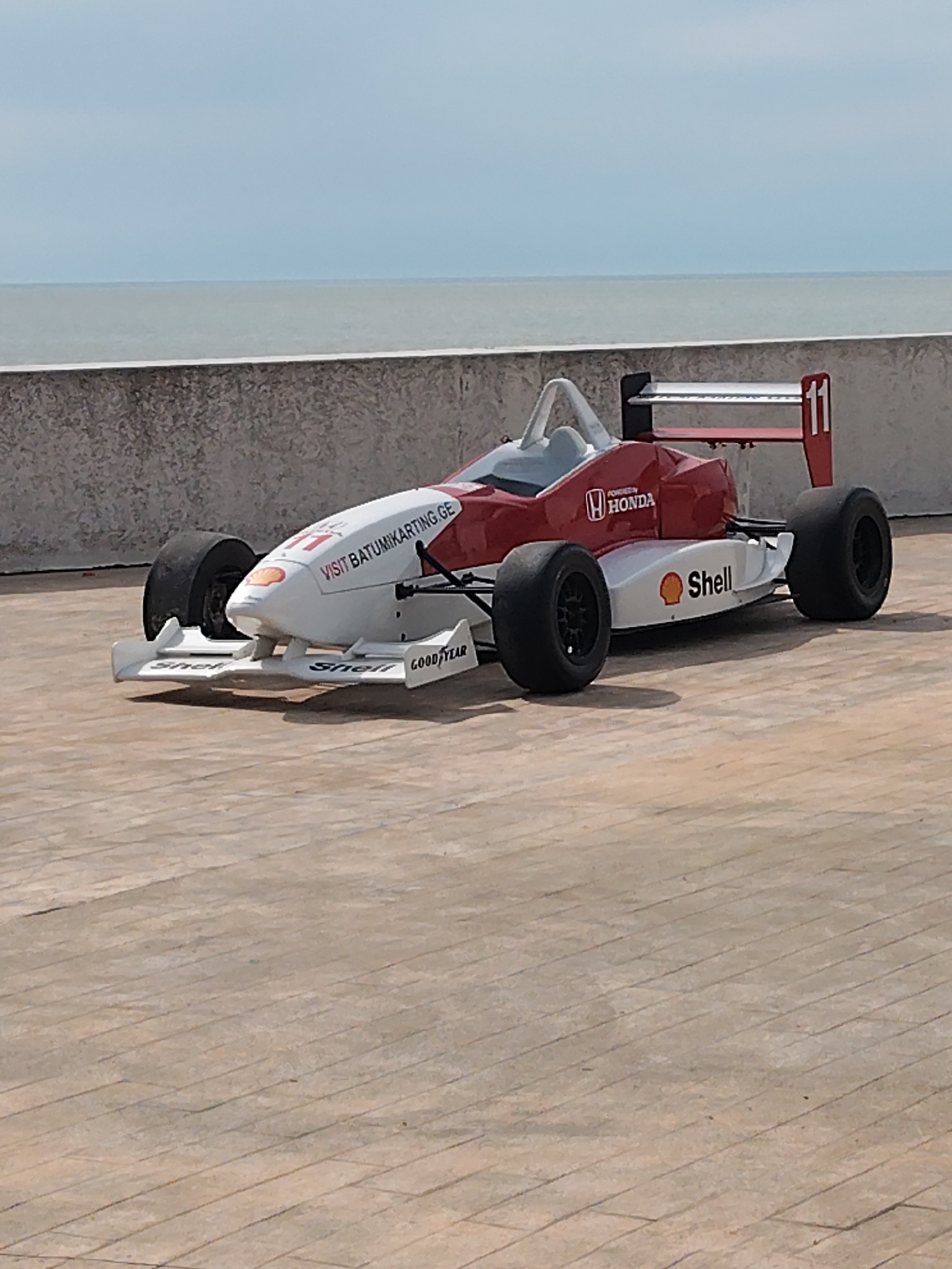 A red and white Formula 1 style go-kart with racing livery bearing the number "11" is parked on a light brown, patterned concrete surface. The kart features logos from Honda, Shell, and Goodyear, along with "VISIT BATUMIKARTING.GE" printed on its side. Behind the kart, there is a low, mottled grey concrete wall, and beyond that, a calm body of water under a light sky. The scene is most likely in the city of Batumi, Georgia. There are no people visible in the image.