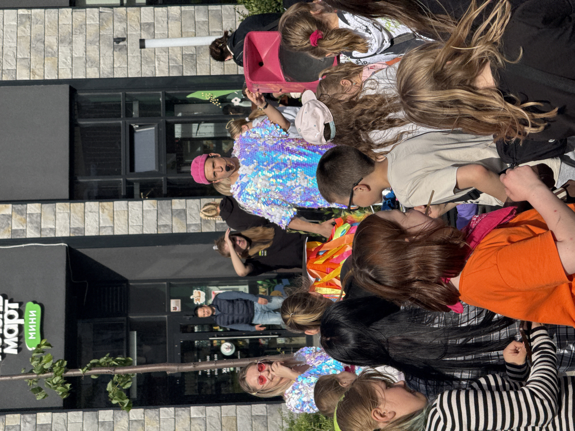 A crowd of children, primarily girls, surround a person wearing a sequined shirt and a pink hat. The person in the sequined shirt appears to be engaging the children, holding a pink megaphone. In the background, the sign "MART MINI" can be seen on a building in what appears to be the city of Yekaterinburg, Russia. Another person is visible standing in a doorway. A tree branch appears in the lower left corner.